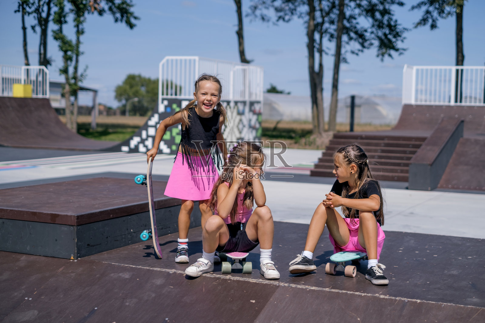 带着滑板的搞笑女孩们坐在滑板公园的金字塔形游乐箱上照片摄影图片