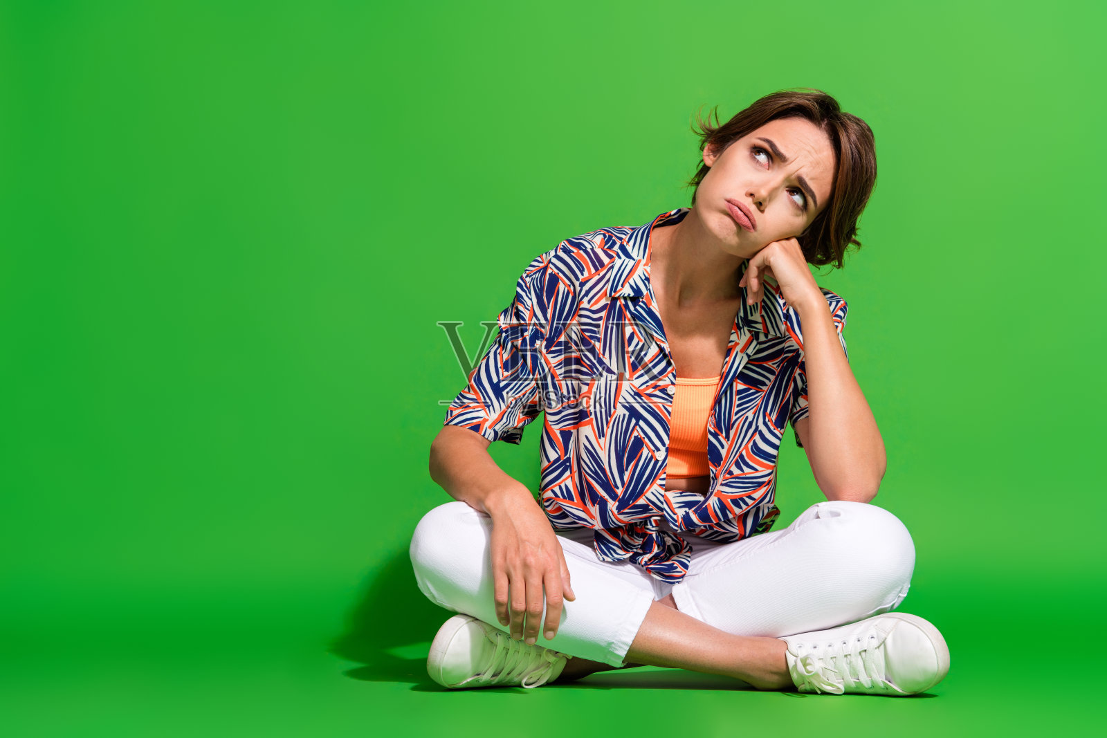 沮丧女子穿印花衬衫，双腿交叉坐着，绿色背景全身照照片摄影图片