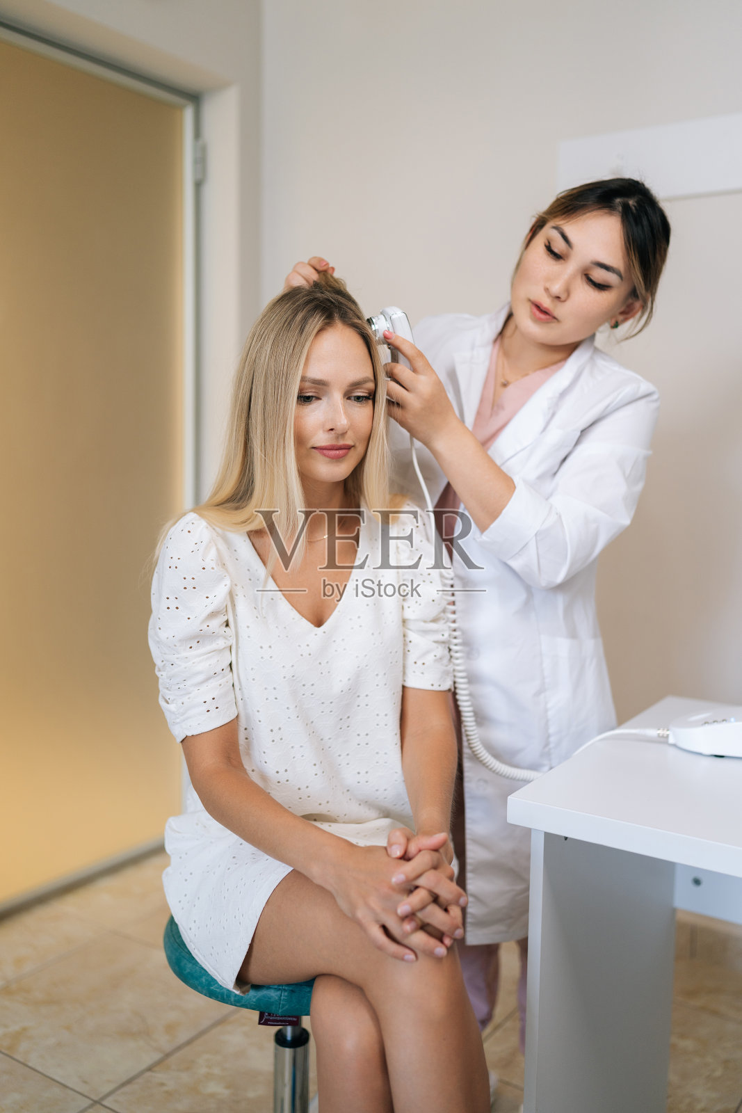 女性毛发医生在皮肤科诊所用专用装置毛发镜诊断金发女性头发的结构和状况的垂直肖像。照片摄影图片