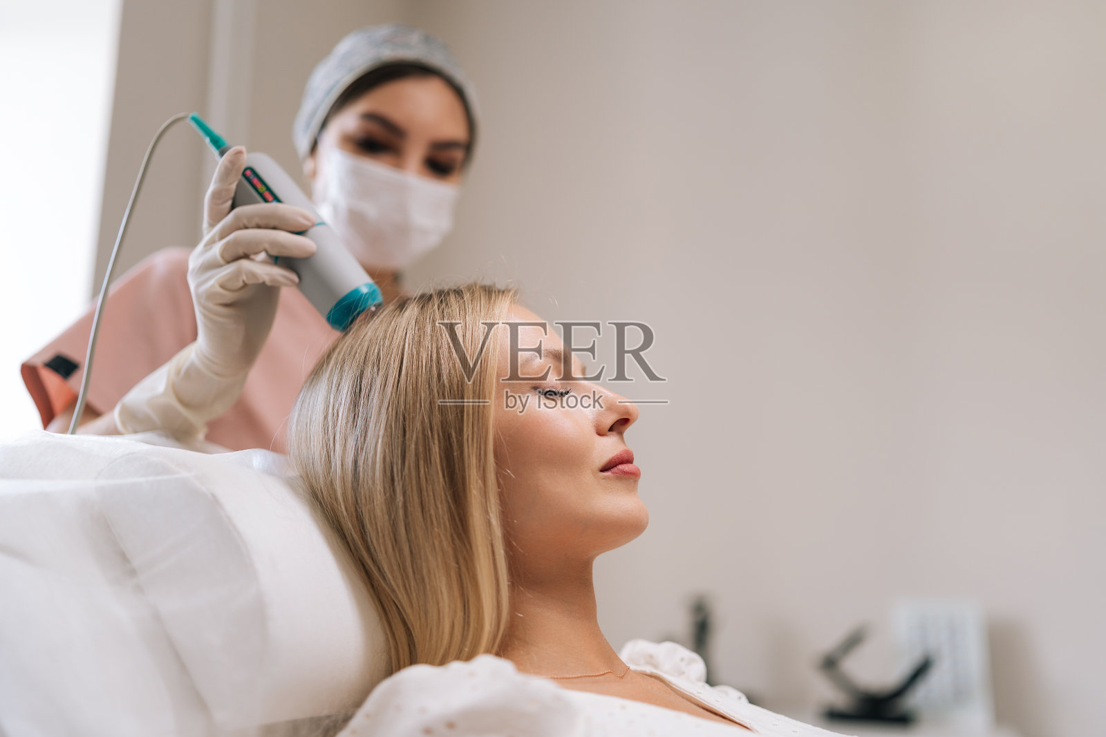 用现代创新仪器进行头发光疗的迷人金发女病人的特写侧视图。毛发学家正在进行头发处理程序照片摄影图片