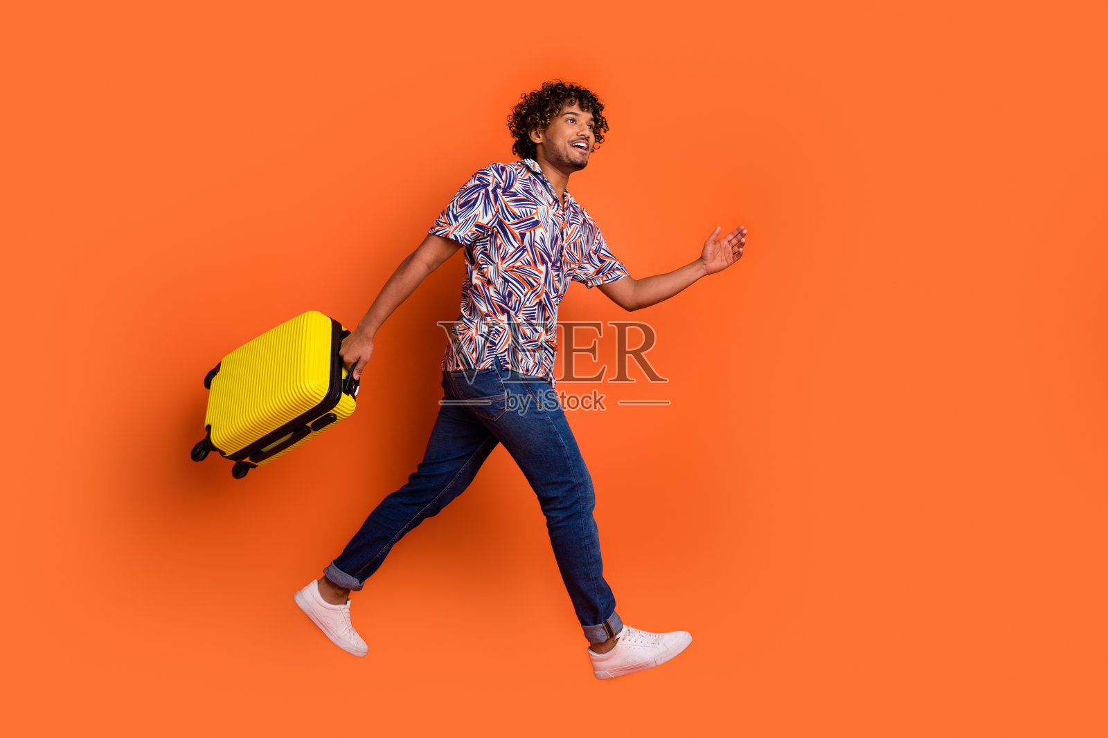 有魅力的年轻人抱着行李跑着的全身照片，他穿着时髦的彩色衣服，孤立在橙色的背景上照片摄影图片