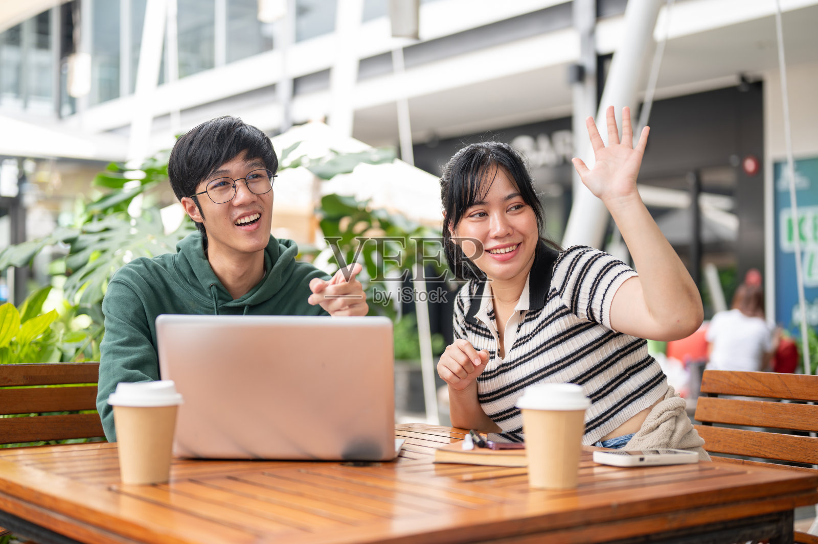 两个兴高采烈的亚洲朋友指着并挥手问候他们遇到的朋友。照片摄影图片