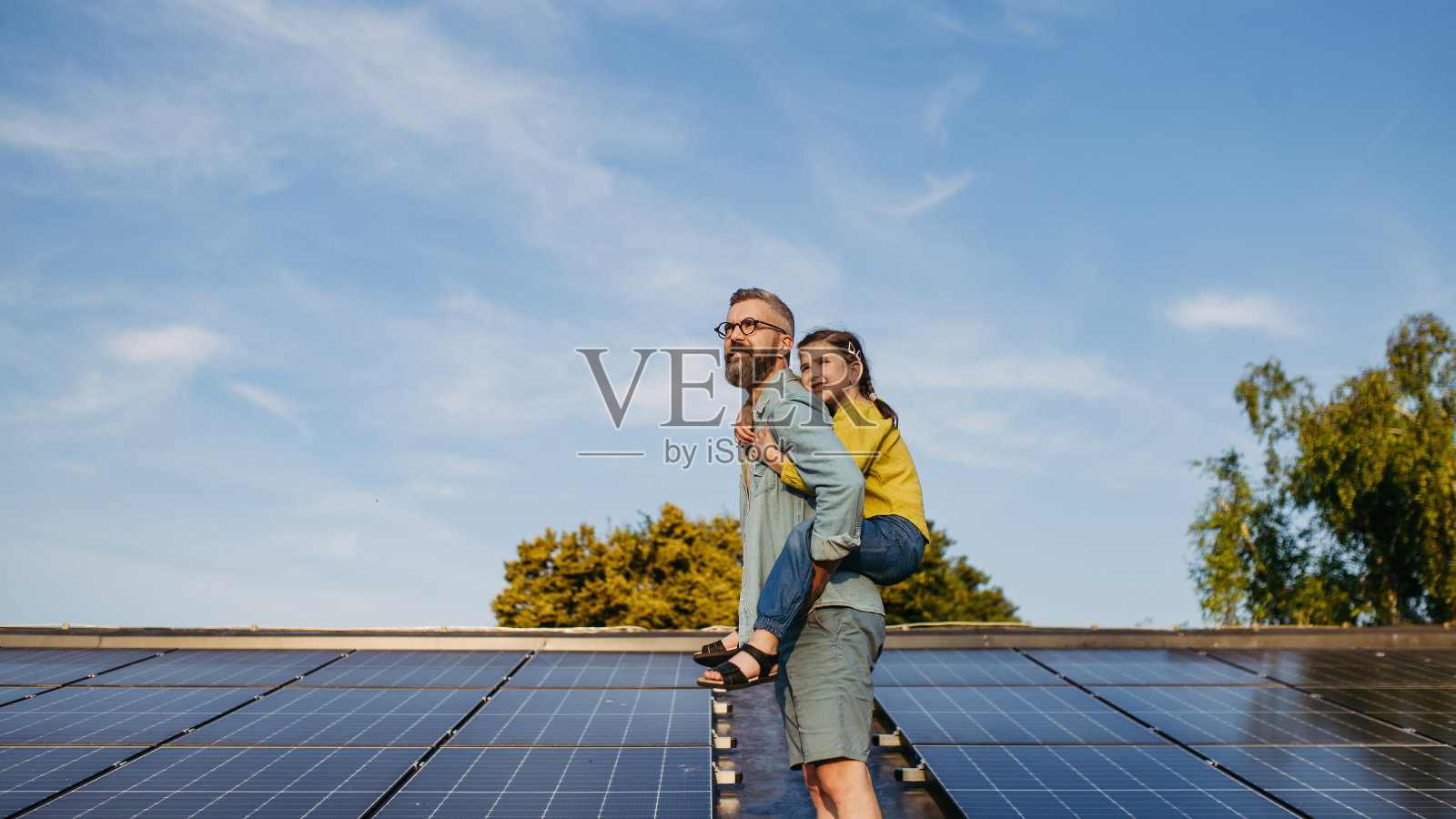 父亲带着女儿坐在屋顶上，背着太阳能板。屋顶太阳能或光伏系统。为下一代创造可持续的未来。照片摄影图片