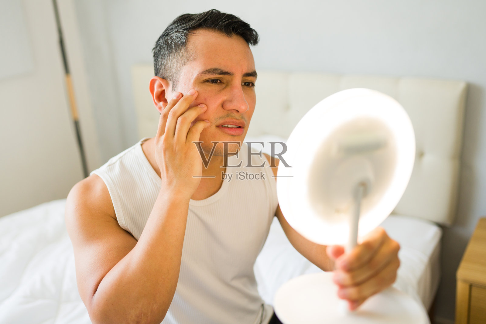 一个有皮肤问题的年轻人摸着自己的脸，凝视着卧室里的一面led镜子照片摄影图片