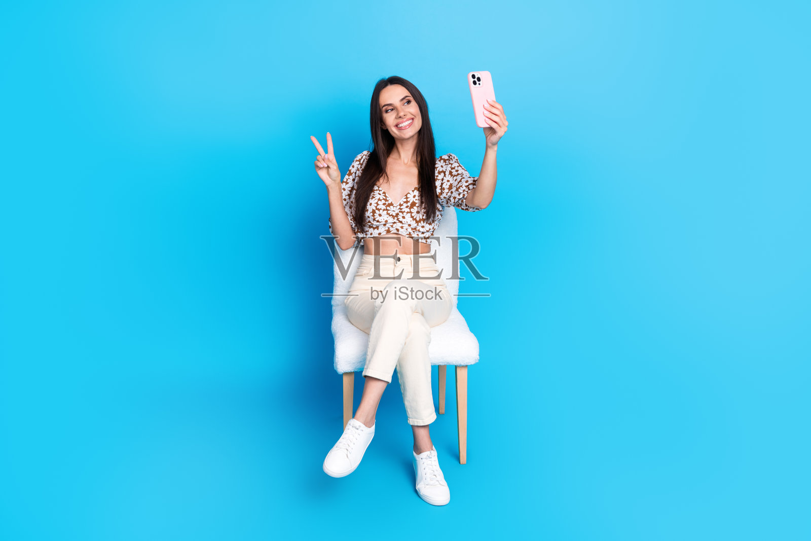 漂亮的年轻女子手机自拍的全尺寸照片v-sign穿着上衣孤立的蓝色背景照片摄影图片
