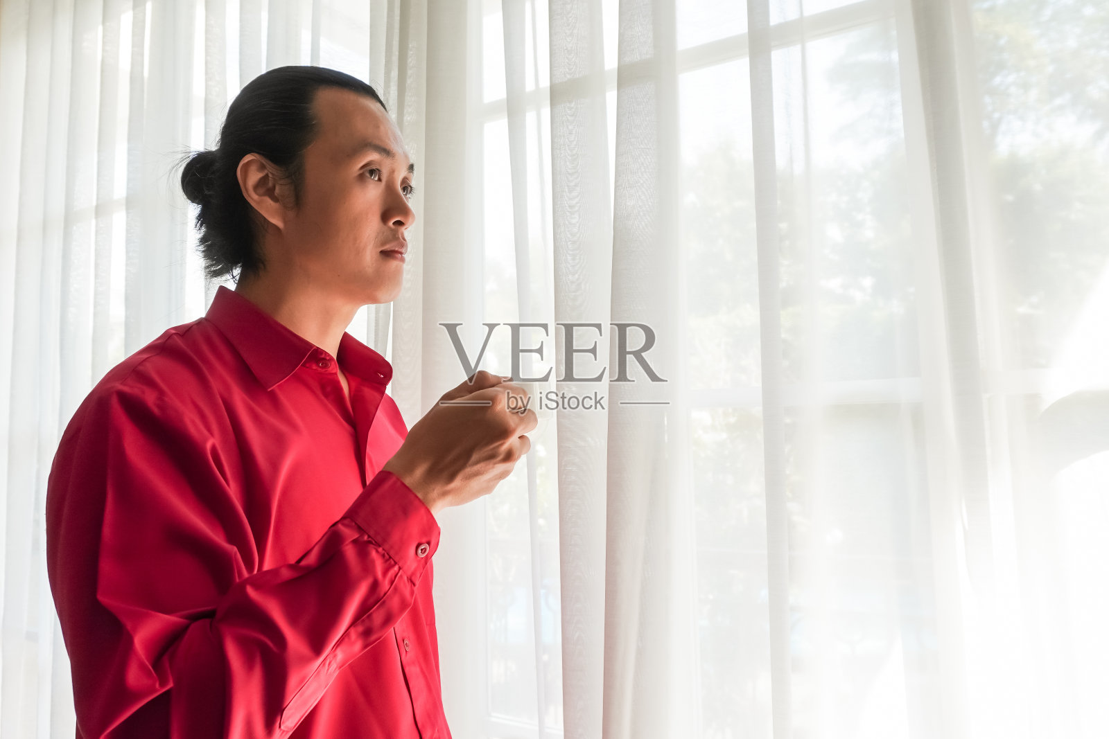 穿着红色长袖衬衫、留着长发的亚洲青年，站在明亮的大窗前，一边喝着咖啡，一边沉思着。空白复制文本空间。照片摄影图片