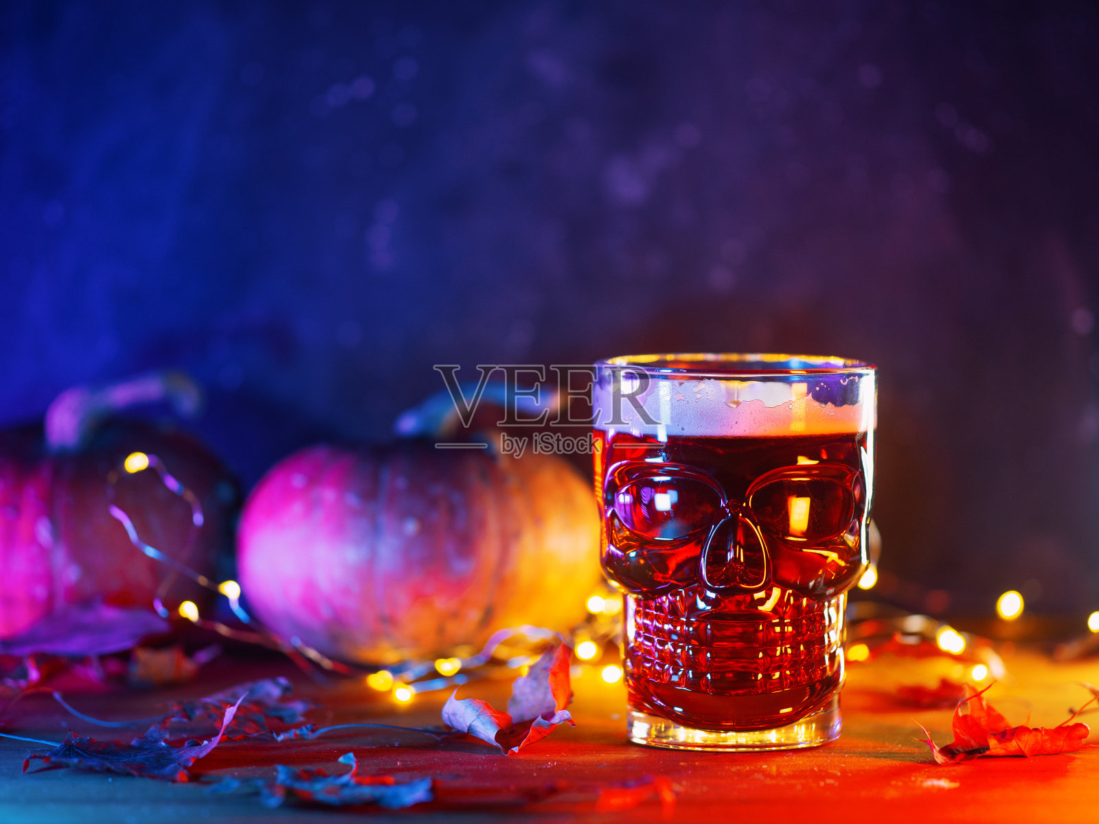 红色麦酒装在一个头骨形状的玻璃杯里，桌子上放着南瓜和秋叶。庆祝万圣节，复制空间照片摄影图片