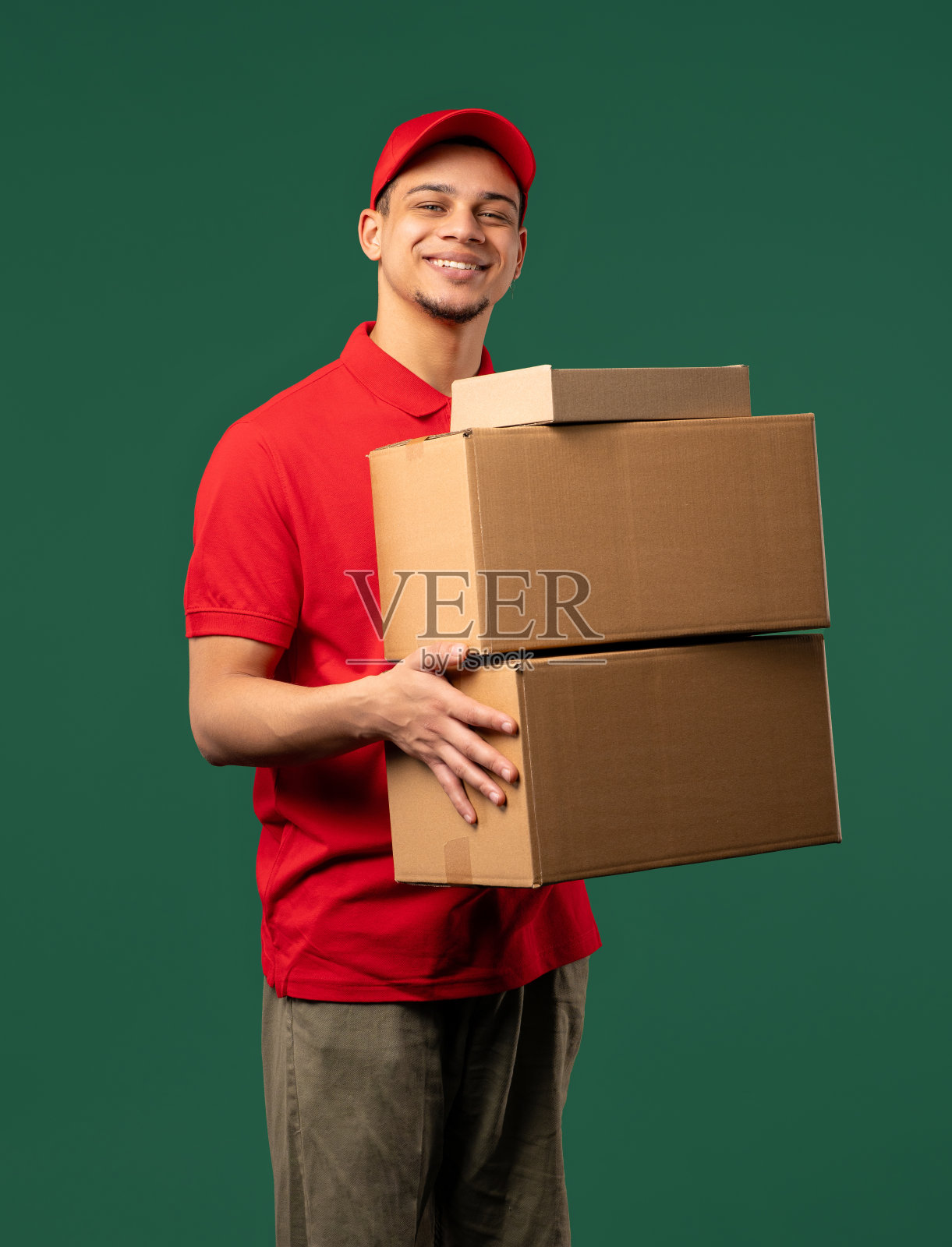 积极的邮政快递员带着包裹箱。快乐的邮差在绿色的工作室背景上投递地址包裹。照片摄影图片