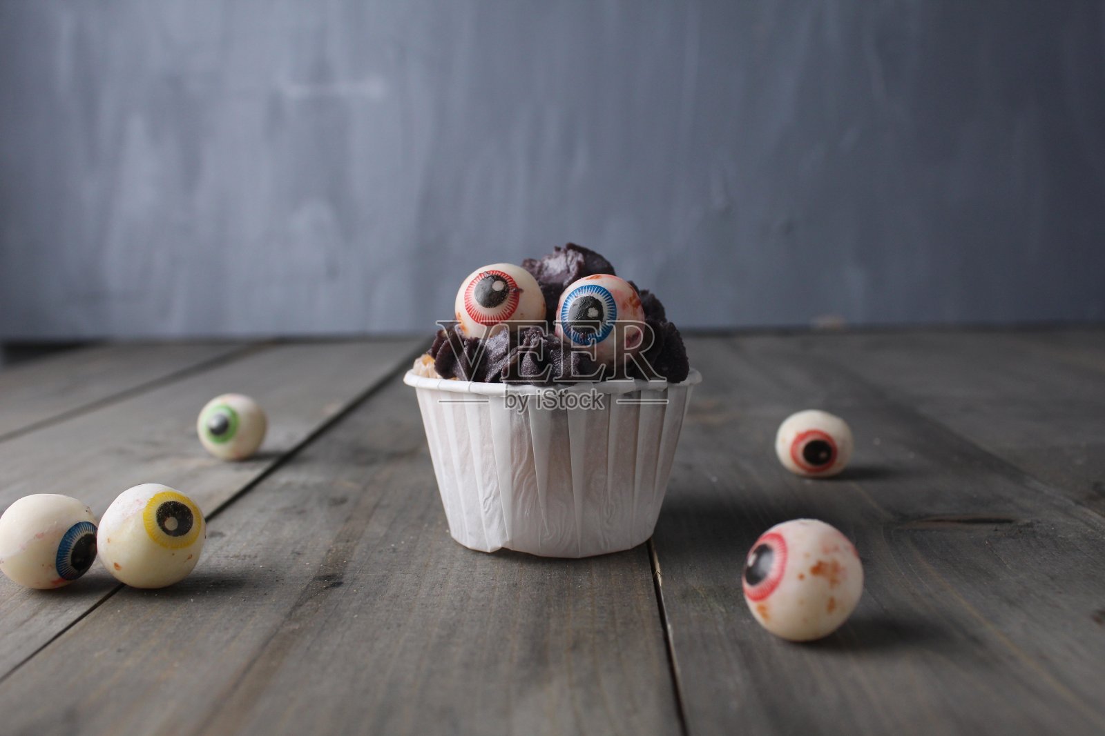 有眼睛的纸杯蛋糕和眼睛形状的软糖并排放在灰色的背景上。万圣节装饰理念照片摄影图片