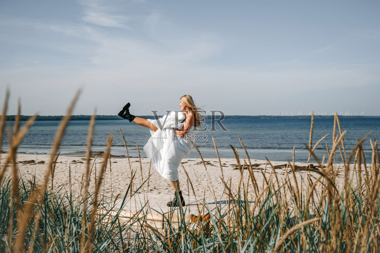 一个年轻女子穿着白色的毯子在沙滩上欢快地跳舞，周围是高高的草地和宁静的大海照片摄影图片