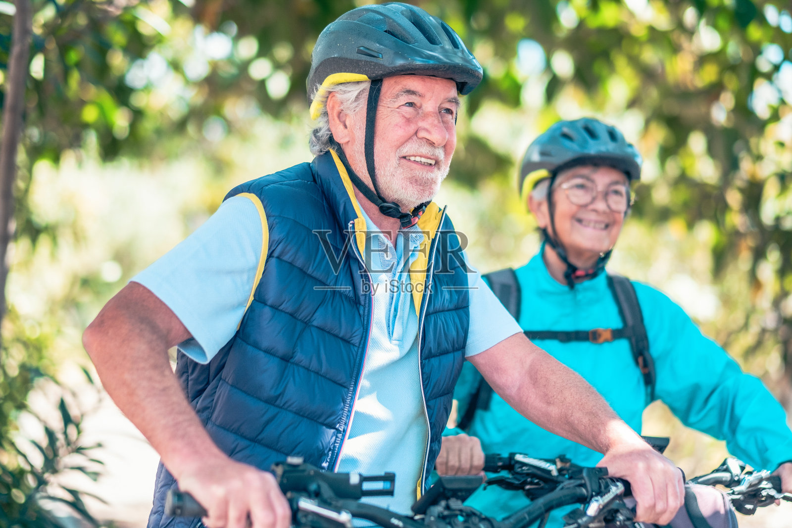 快乐活跃的老年白人夫妇骑着电动自行车，戴着头盔在公园里跑步。微笑的退休老人在大自然中跑步，享受健康的生活方式照片摄影图片