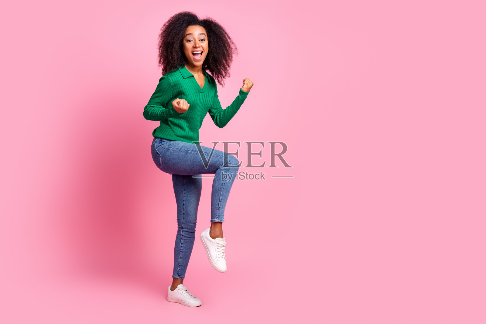 迷人的年轻女子赢得了举起拳头的全身照片，她穿着时髦的绿色衣服，孤立在粉红色的背景上照片摄影图片