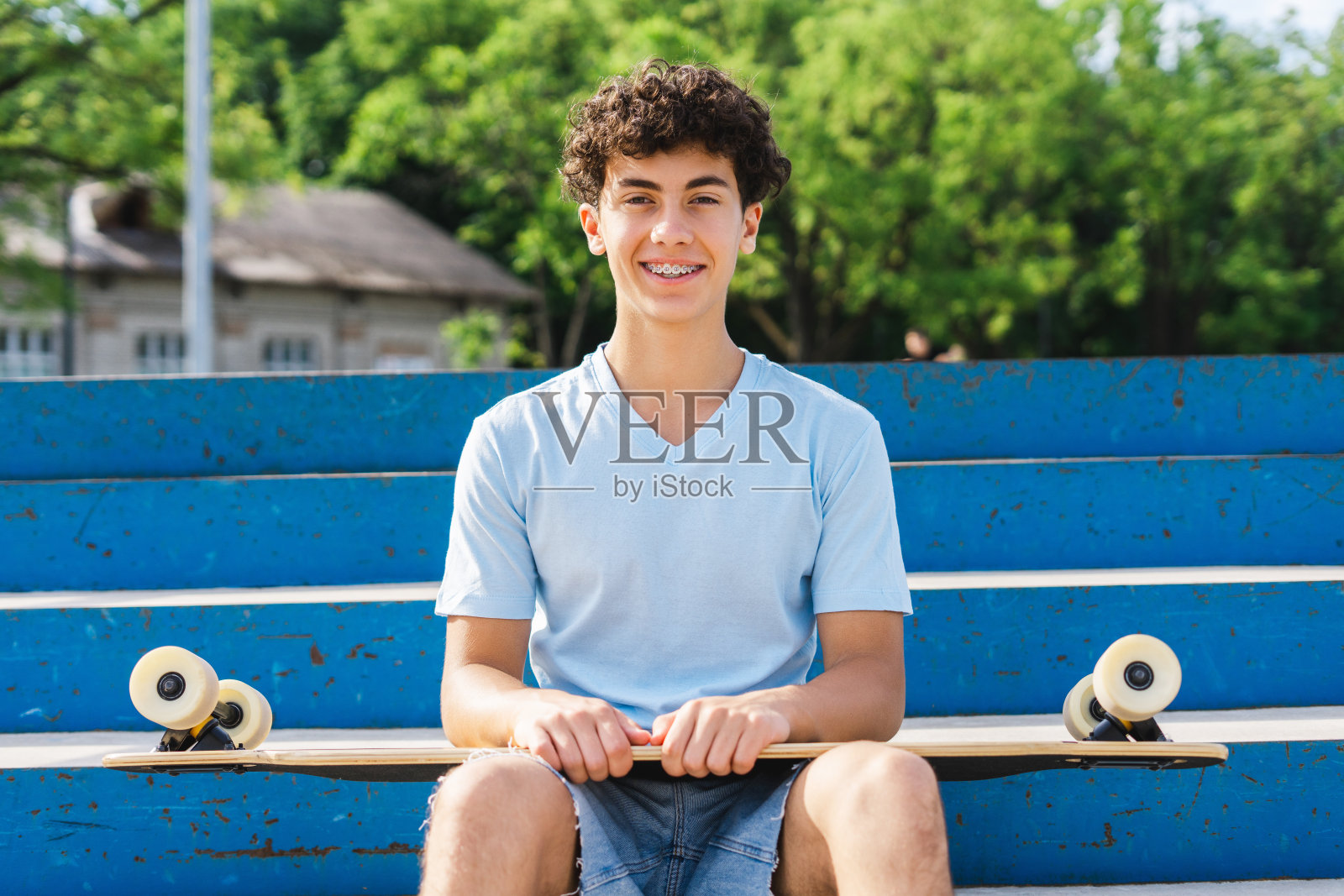 微笑的十几岁的男孩，带着背带，拿着滑板，坐在球场上看着镜头照片摄影图片