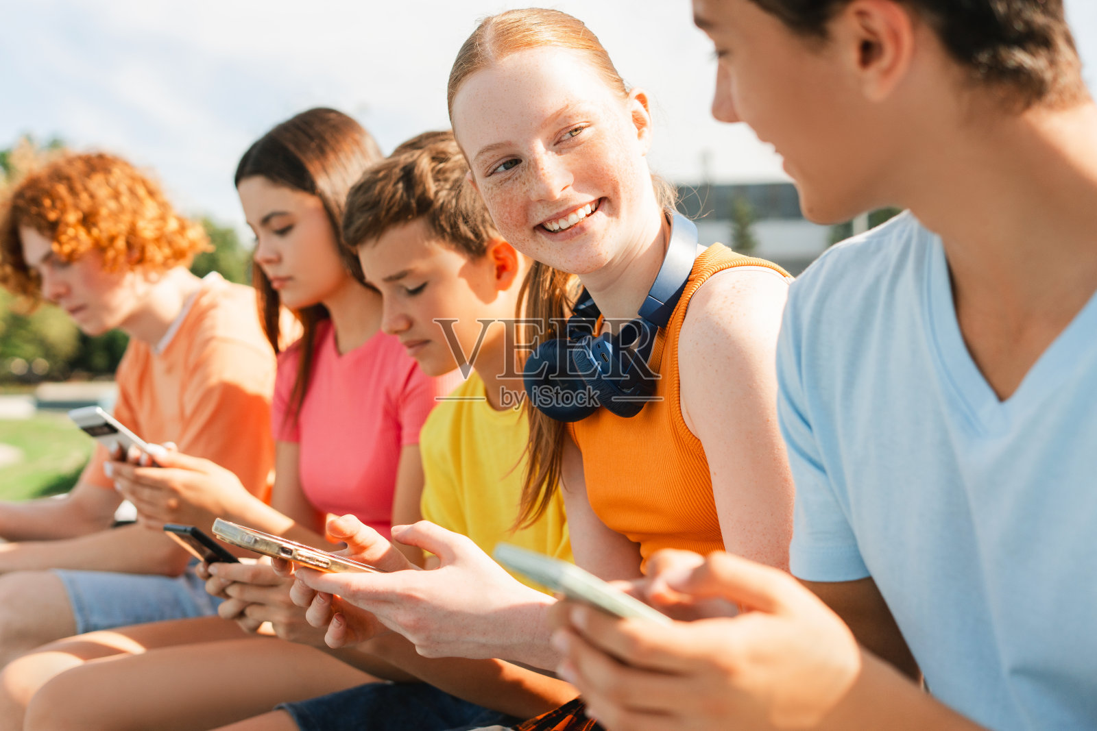 快乐的少女拿着智能手机和耳机和朋友坐在户外照片摄影图片