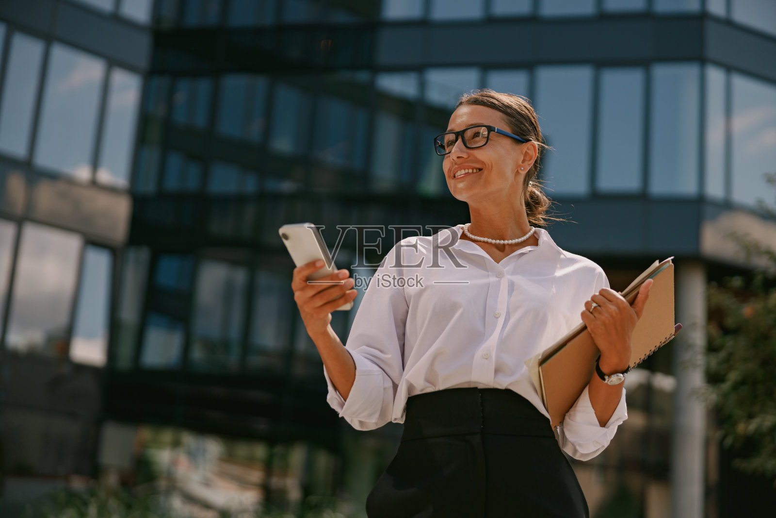 一位自信的女商人，在一栋现代化的办公楼外拿着智能手机和文件夹照片摄影图片