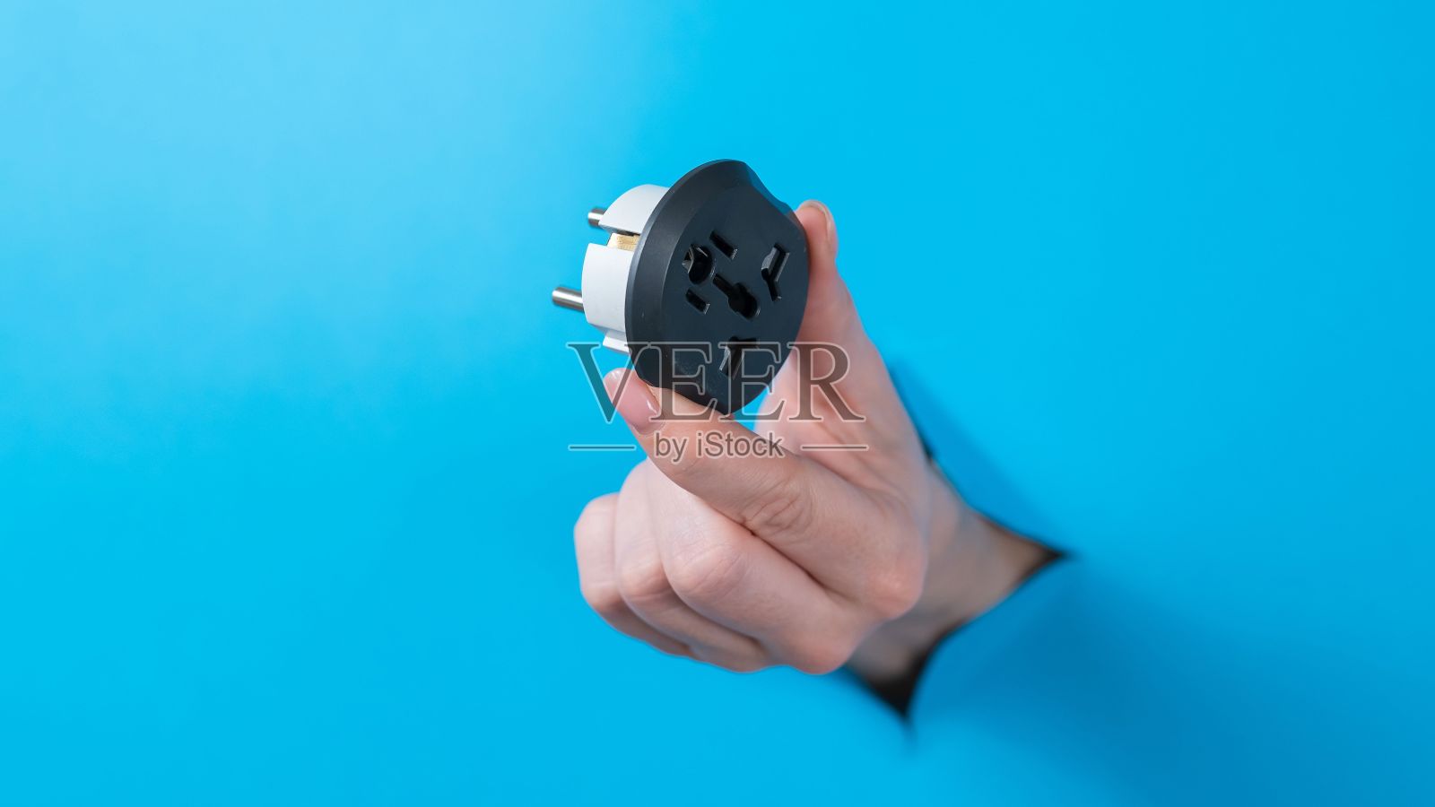 一个女人拿着插座适配器。一个女人的手从蓝色背景的一个洞里伸出来。照片摄影图片