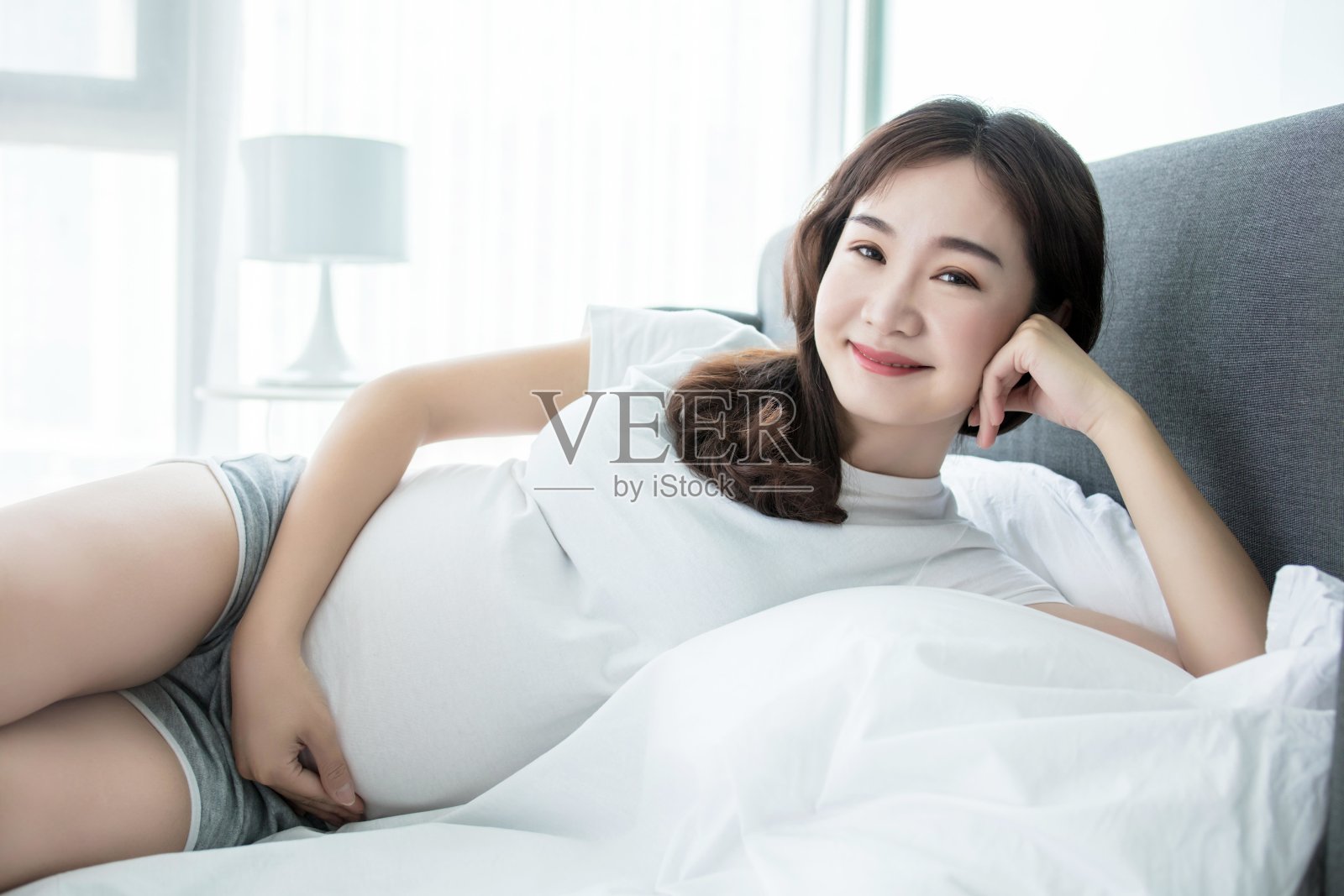 一位孕妇穿着白衬衫和灰色短裤躺在床上，手放在肚子上，面带微笑照片摄影图片