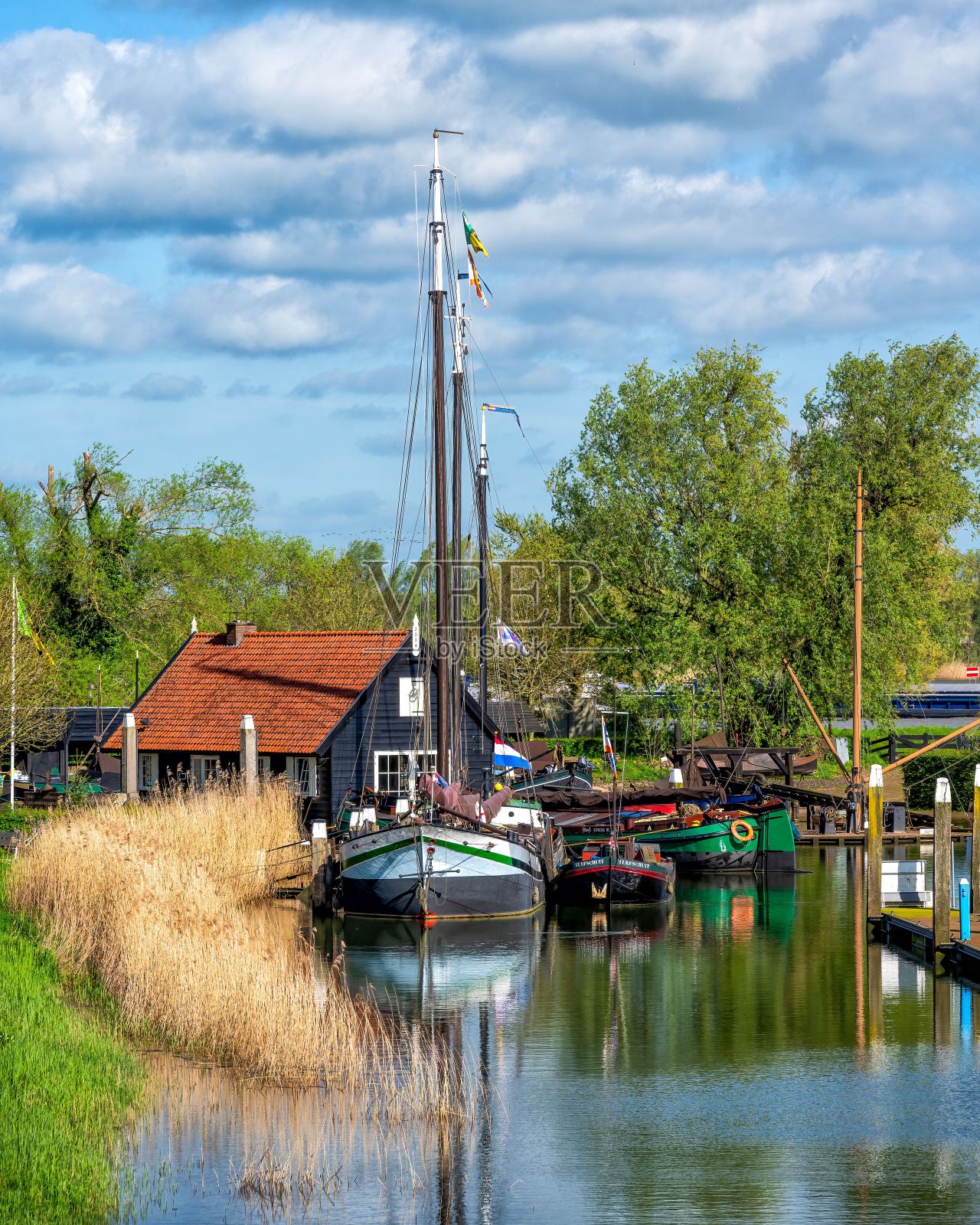 在多云的天空下，小船停靠在有小房子和树木的运河旁。照片摄影图片