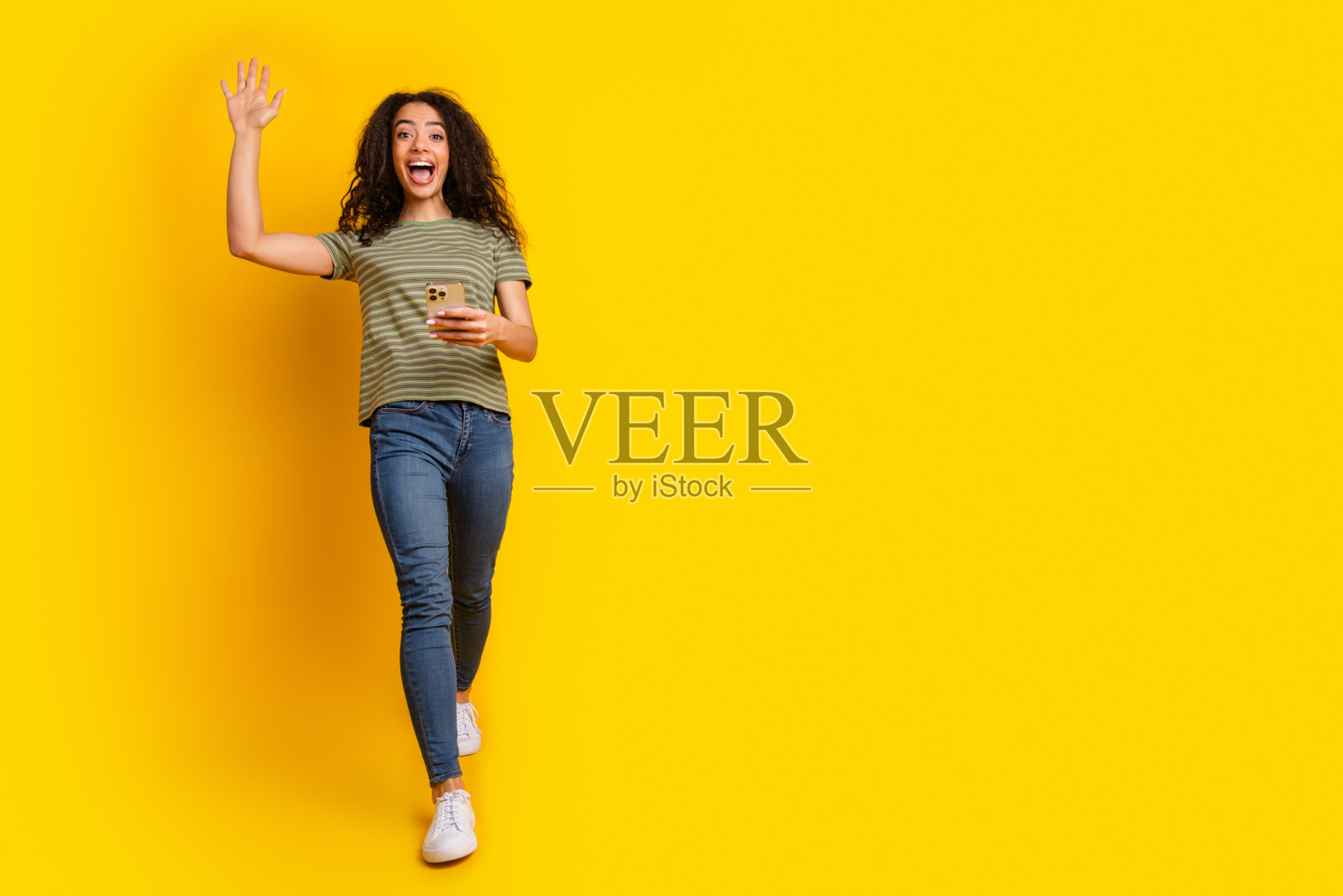 穿着t恤的可爱漂亮女孩的全尺寸照片，看着智能手机挥舞着手掌，靠近黄色背景的空白区域照片摄影图片