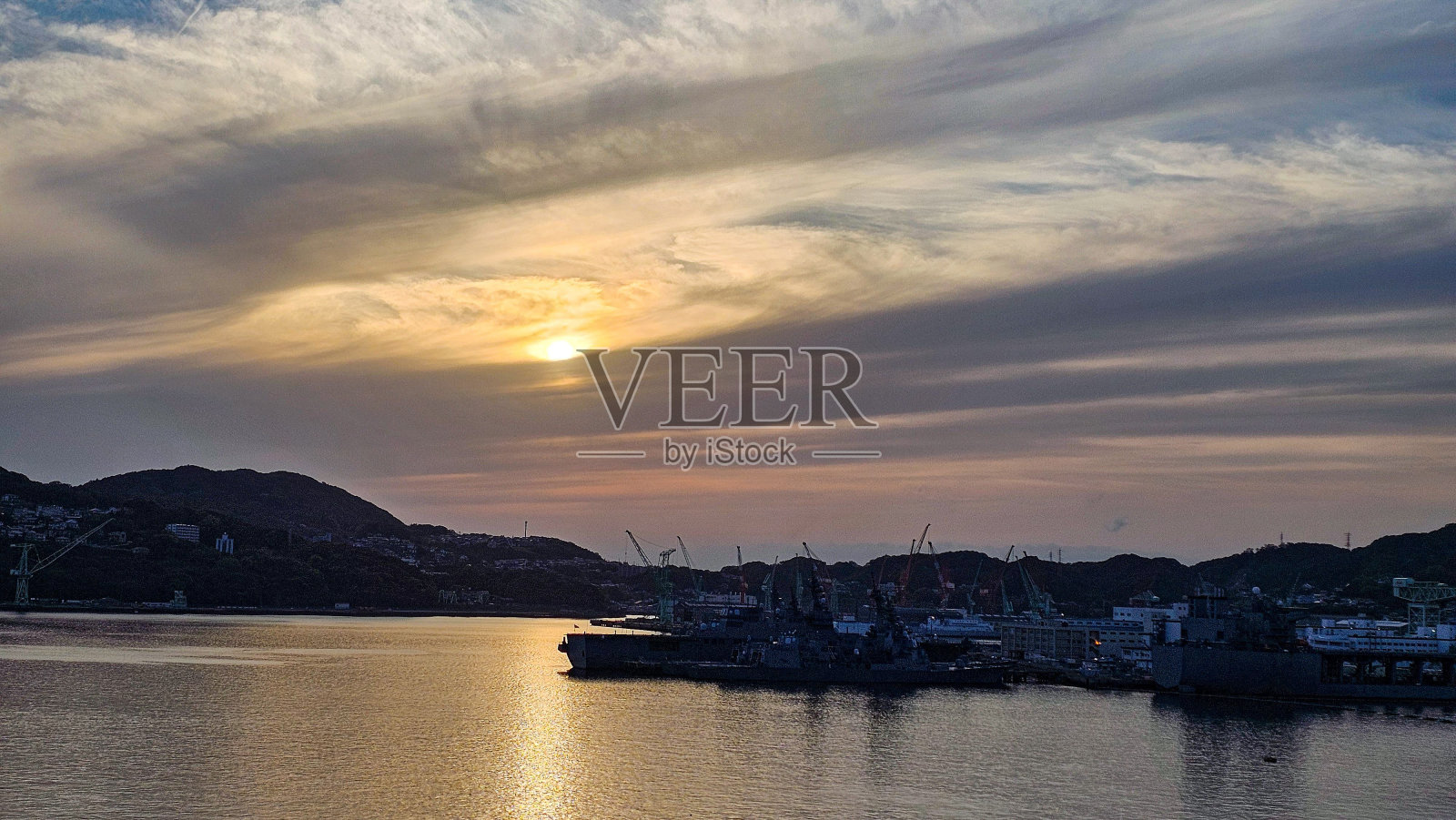 在长崎佐世保，宁静的落日映照着海港上的船只和起重机，映照着平静的水面照片摄影图片