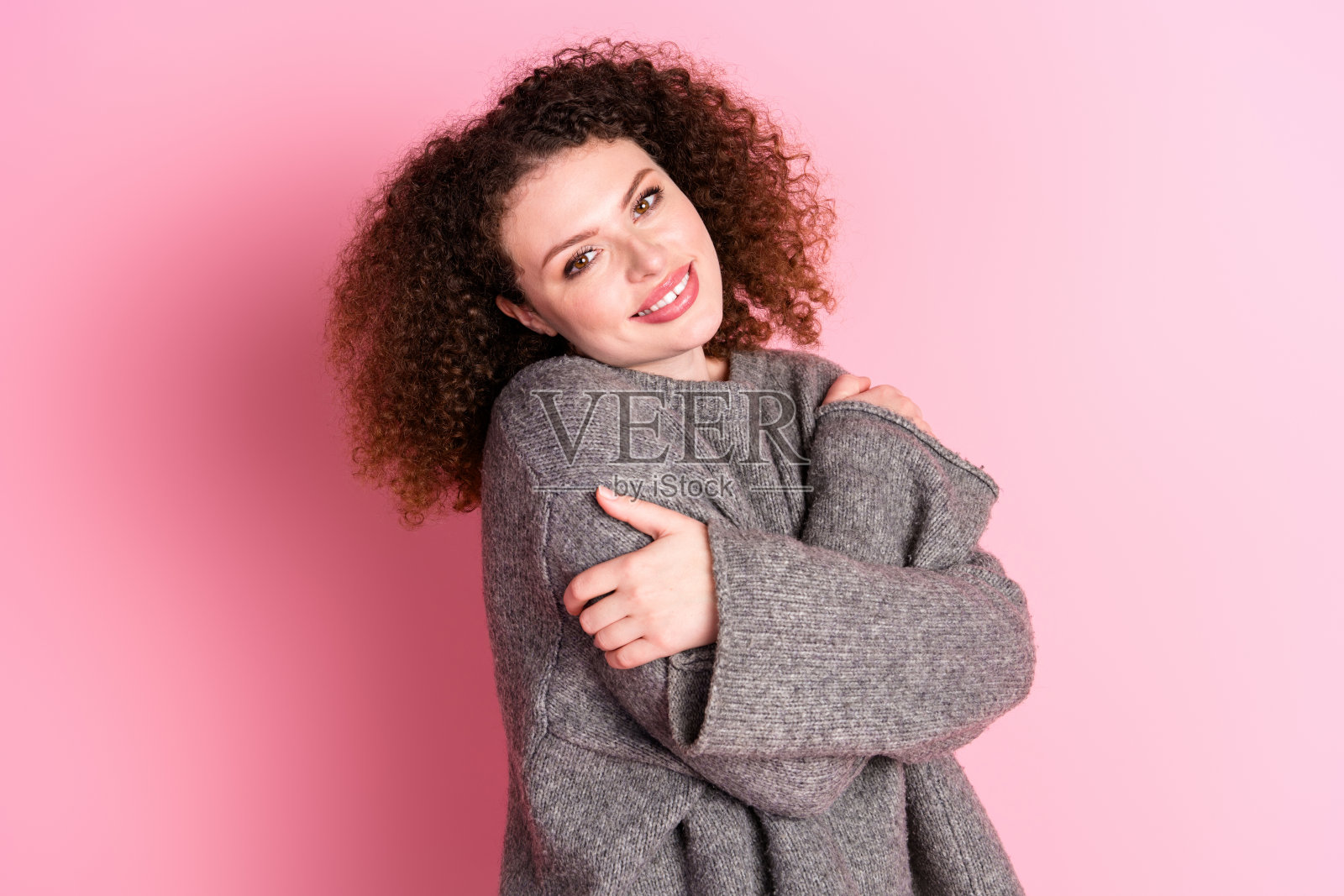 美丽的年轻女孩拥抱自己，享受温暖，穿着时髦的灰色服装，孤立在粉红色的背景照片摄影图片