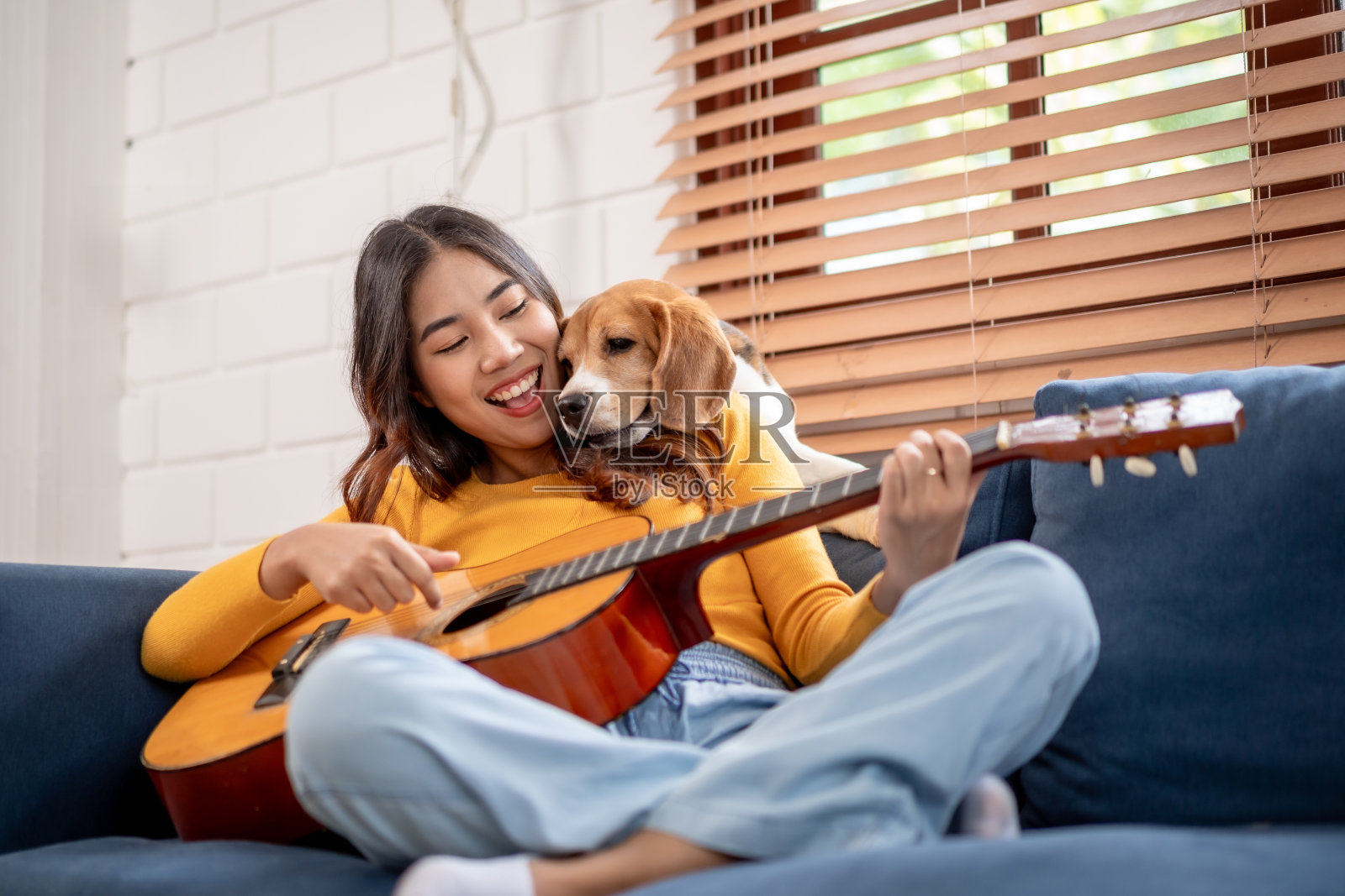 亚洲美女在家里的客厅里和她的小猎犬玩耍。年轻的女主人心情愉快，放松，享受吉他和她可爱的宠物小狗一起在家里度过闲暇时光照片摄影图片