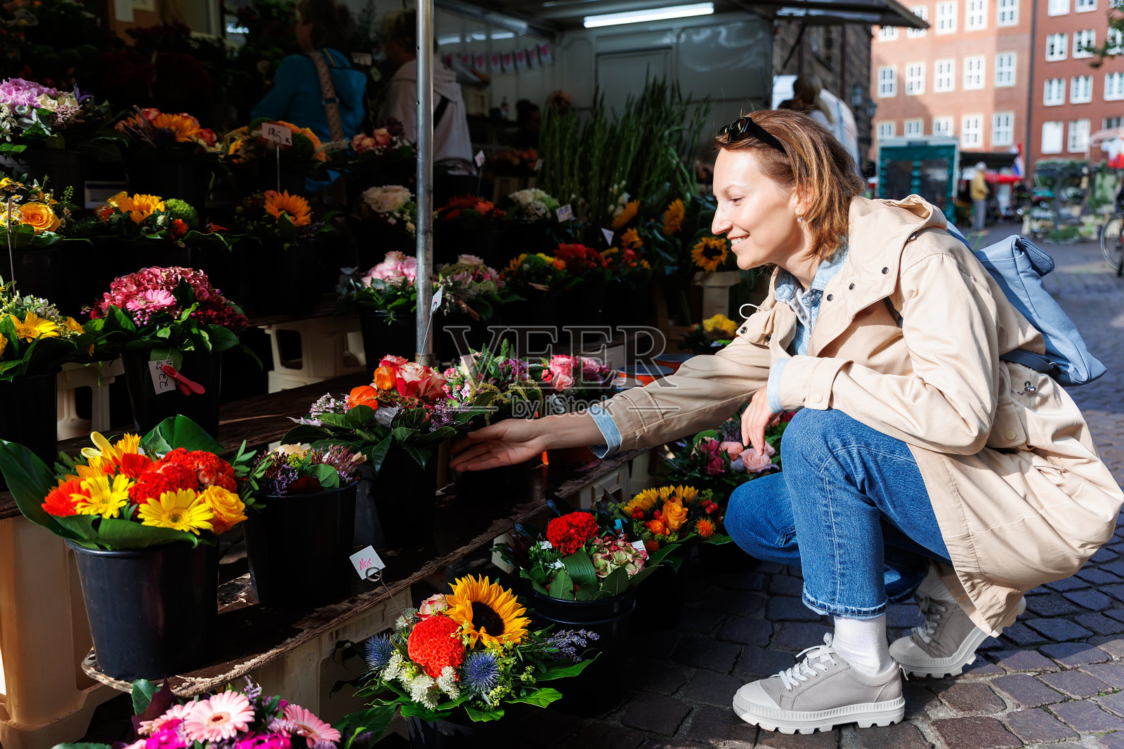 年轻的成年快乐的女子跪在户外市场的摊位上选择购买五颜六色的盆花。在阳光明媚的春日，德国植物街商店出售盆栽开花植物照片摄影图片