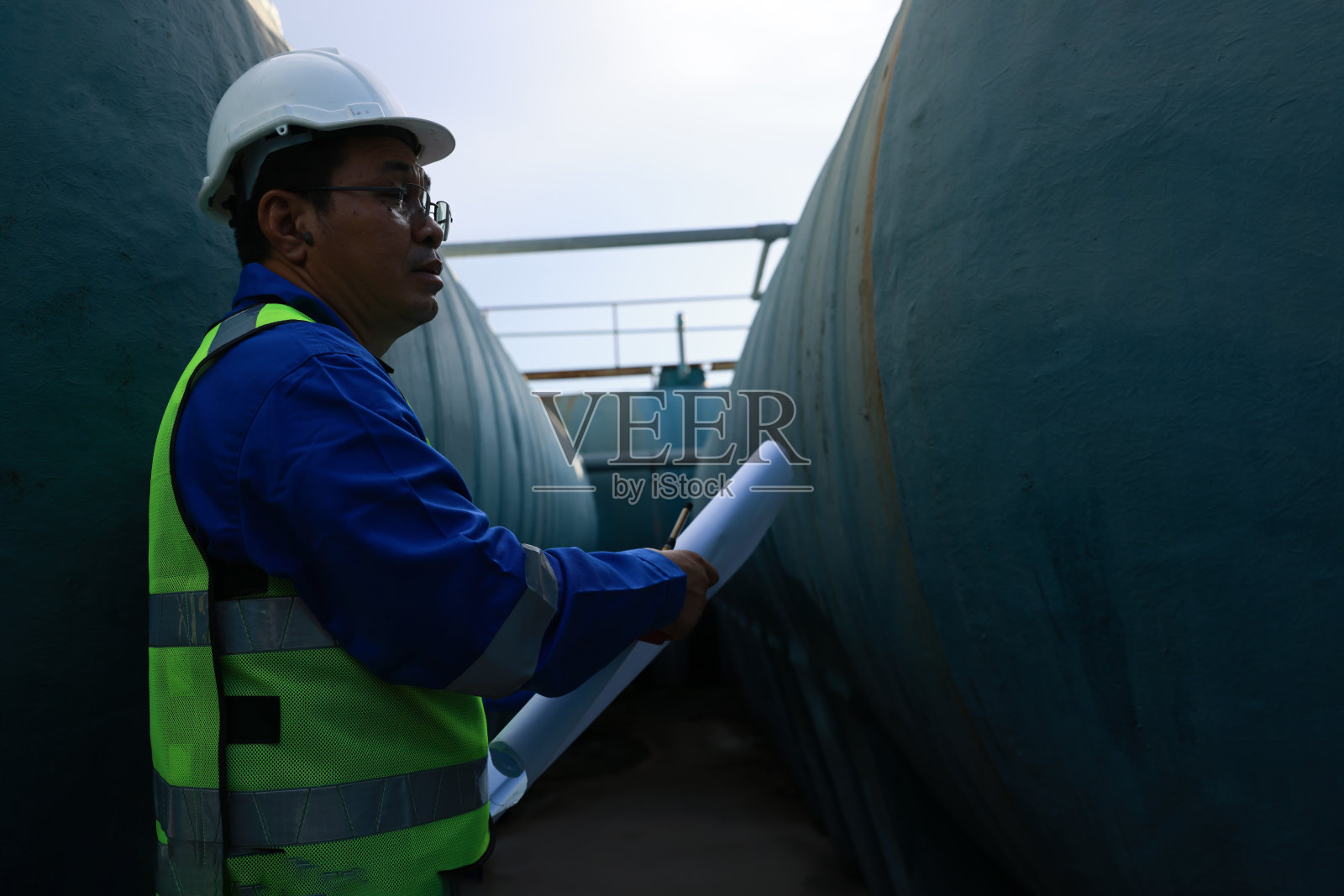 身穿反光背心，手持对讲机，在工厂施工现场的大型水箱前绘制图纸的高级维修工程师。照片摄影图片