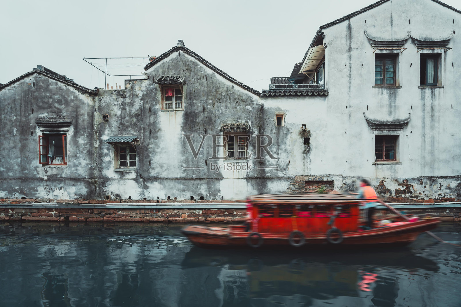 一艘红船驶过平静的运河，经过一排白墙破旧的中国传统建筑。照片摄影图片