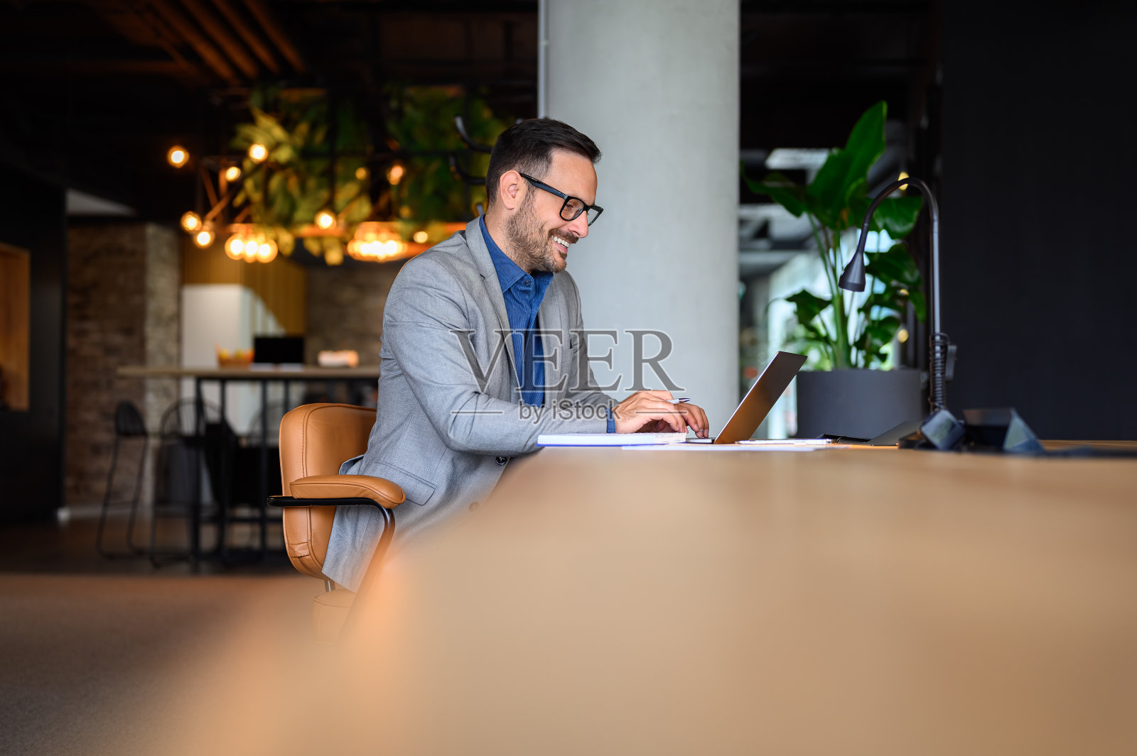 戴着眼镜微笑的年轻商人坐在现代办公室的办公桌前，在笔记本电脑上工作照片摄影图片