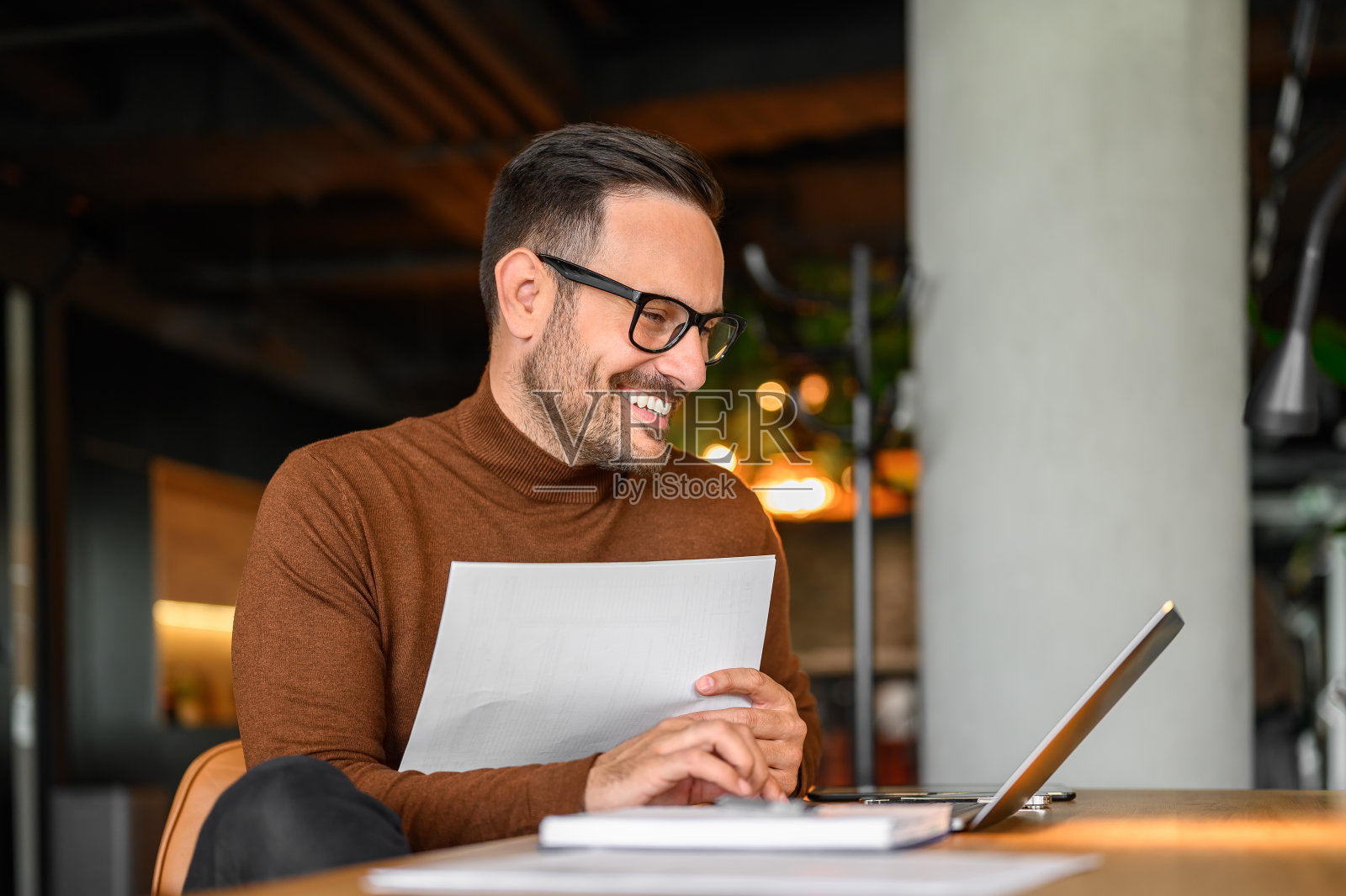 快乐的商人戴着眼镜，拿着文件，在办公室的办公桌上用笔记本电脑检查预算照片摄影图片