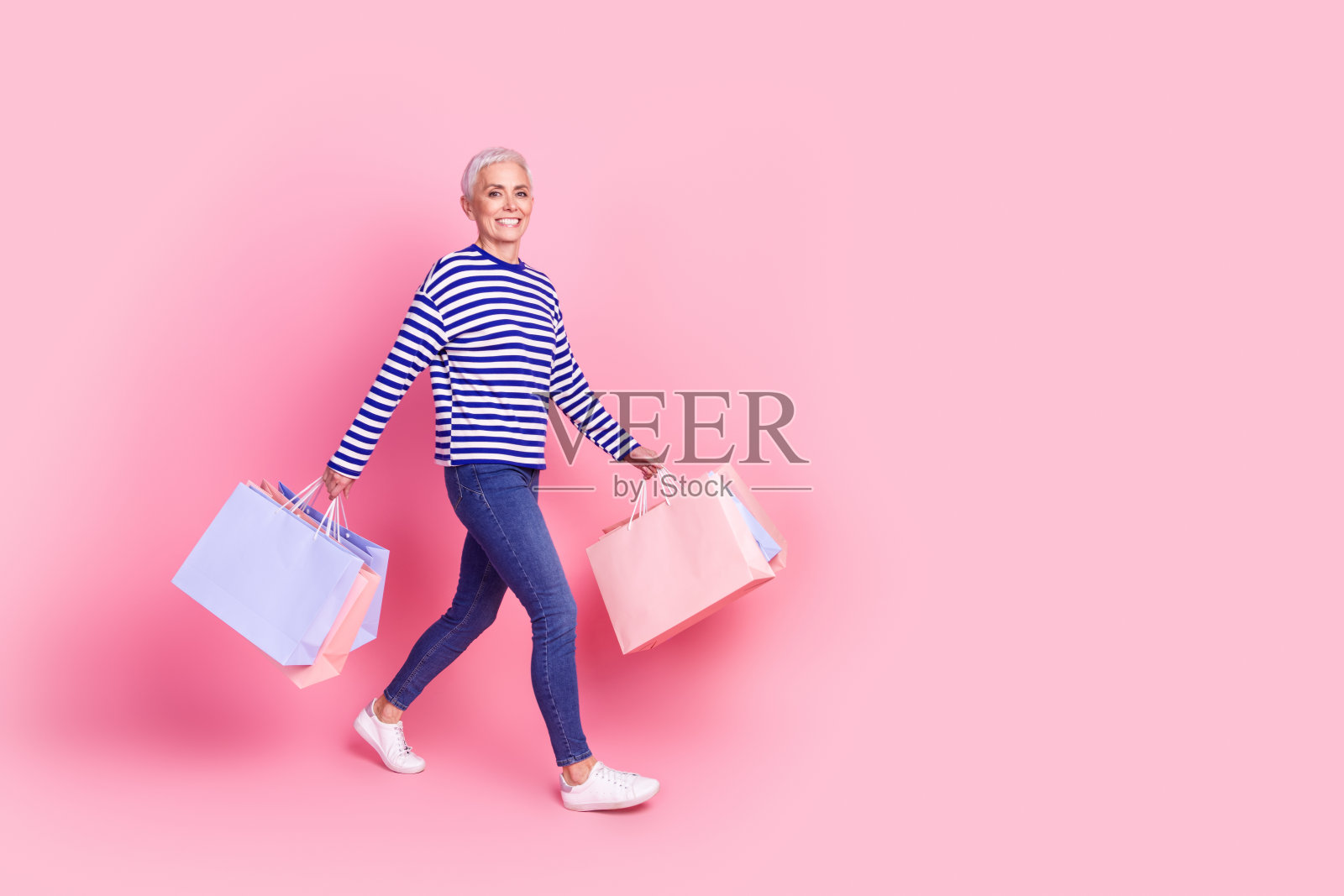 照片中可爱的退休妇女穿着条纹时尚的衣服走在空旷的空间里，在粉红色的背景上背着包包照片摄影图片