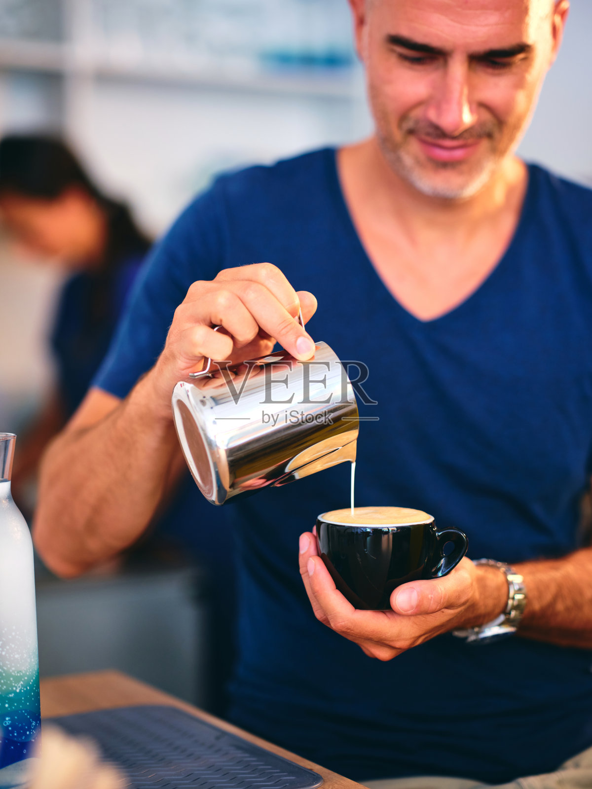 一个穿着蓝色衬衫的男人，脸上带着微笑，艺术地把牛奶从金属壶里倒进一个黑色的咖啡杯里，这是一个极具美感的咖啡馆。照片摄影图片