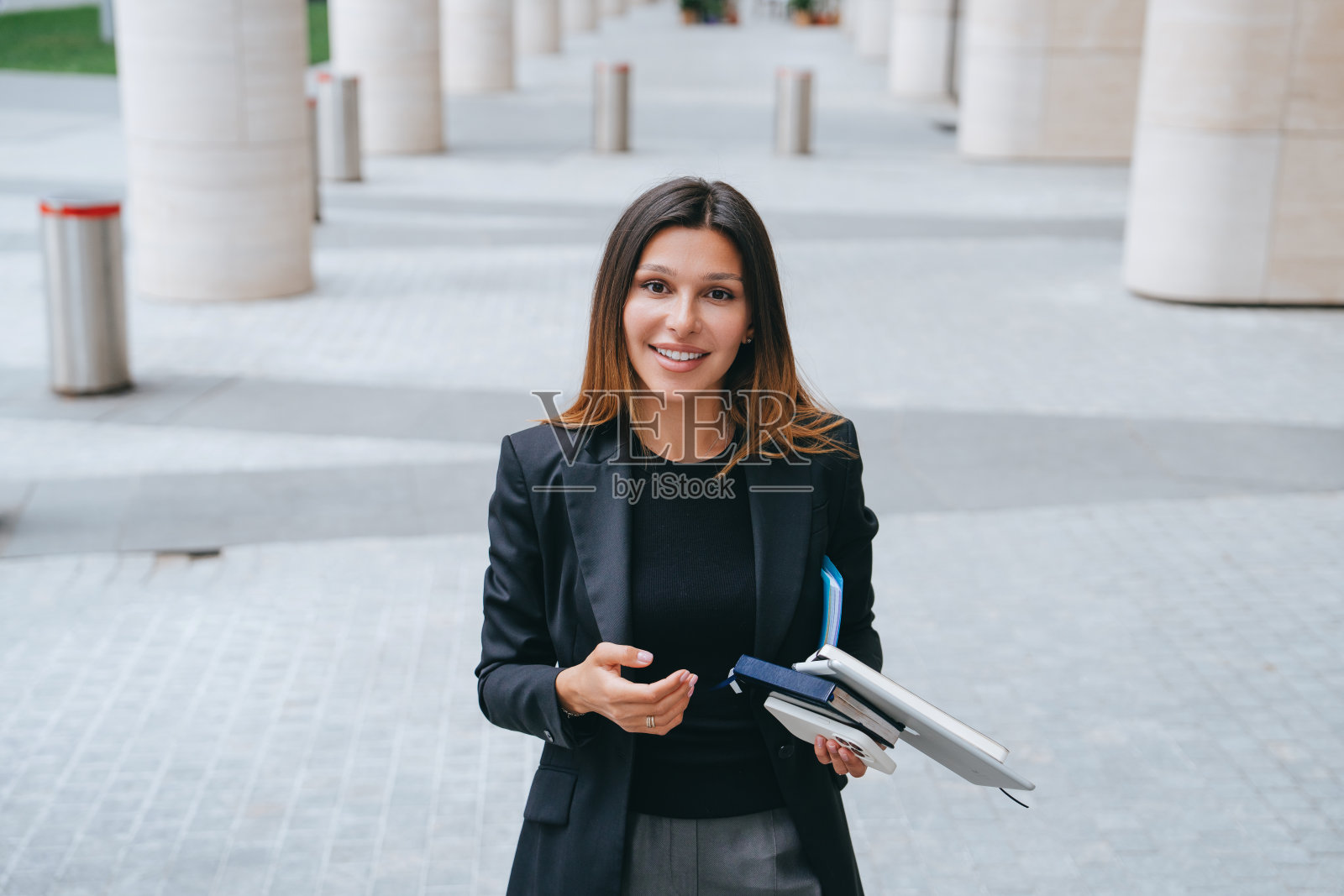 一位身着黑色运动上衣、面带微笑的女商人，手里拿着笔记本和文件，站在有着现代建筑的户外城市环境中照片摄影图片