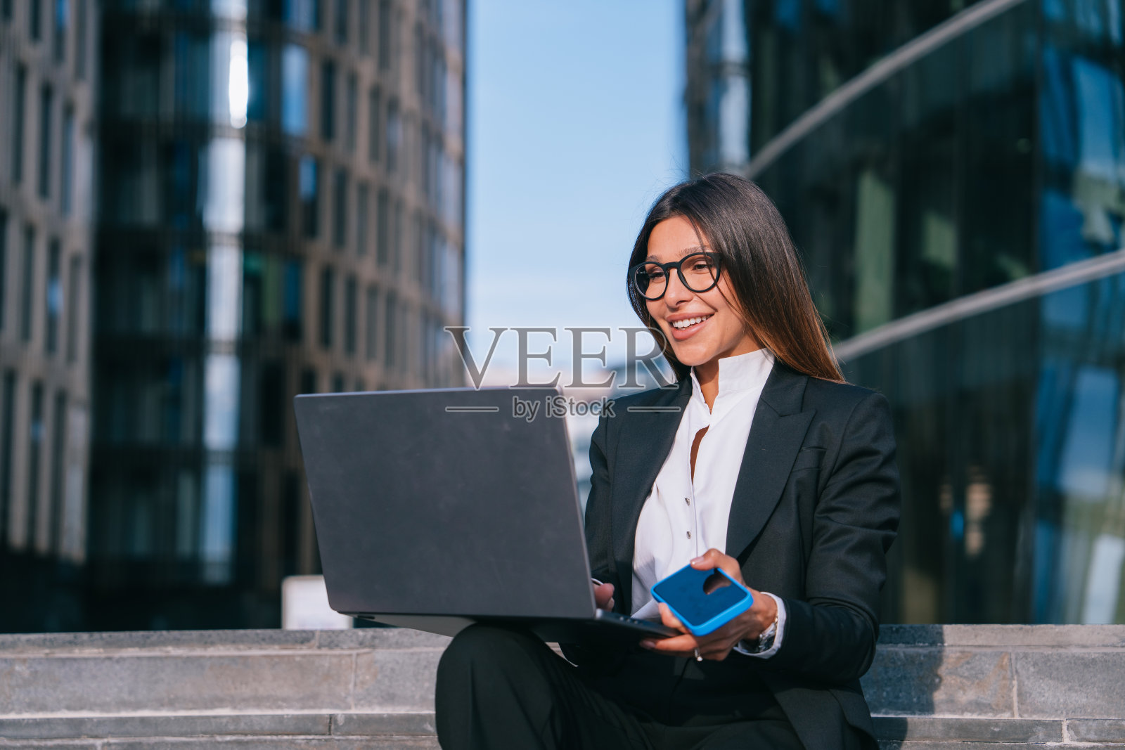 一位身着黑色西装、面带微笑的女商人在户外用笔记本电脑工作，手里拿着一部蓝色智能手机，背景是现代建筑照片摄影图片