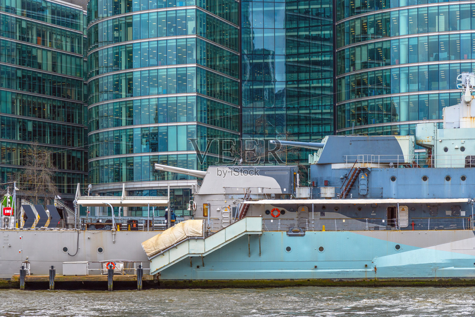 漂浮的战争博物馆船HMS贝尔法斯特的侧视图停靠在泰晤士河上，在多云的天空下，河对面有玻璃封闭的办公楼。照片摄影图片