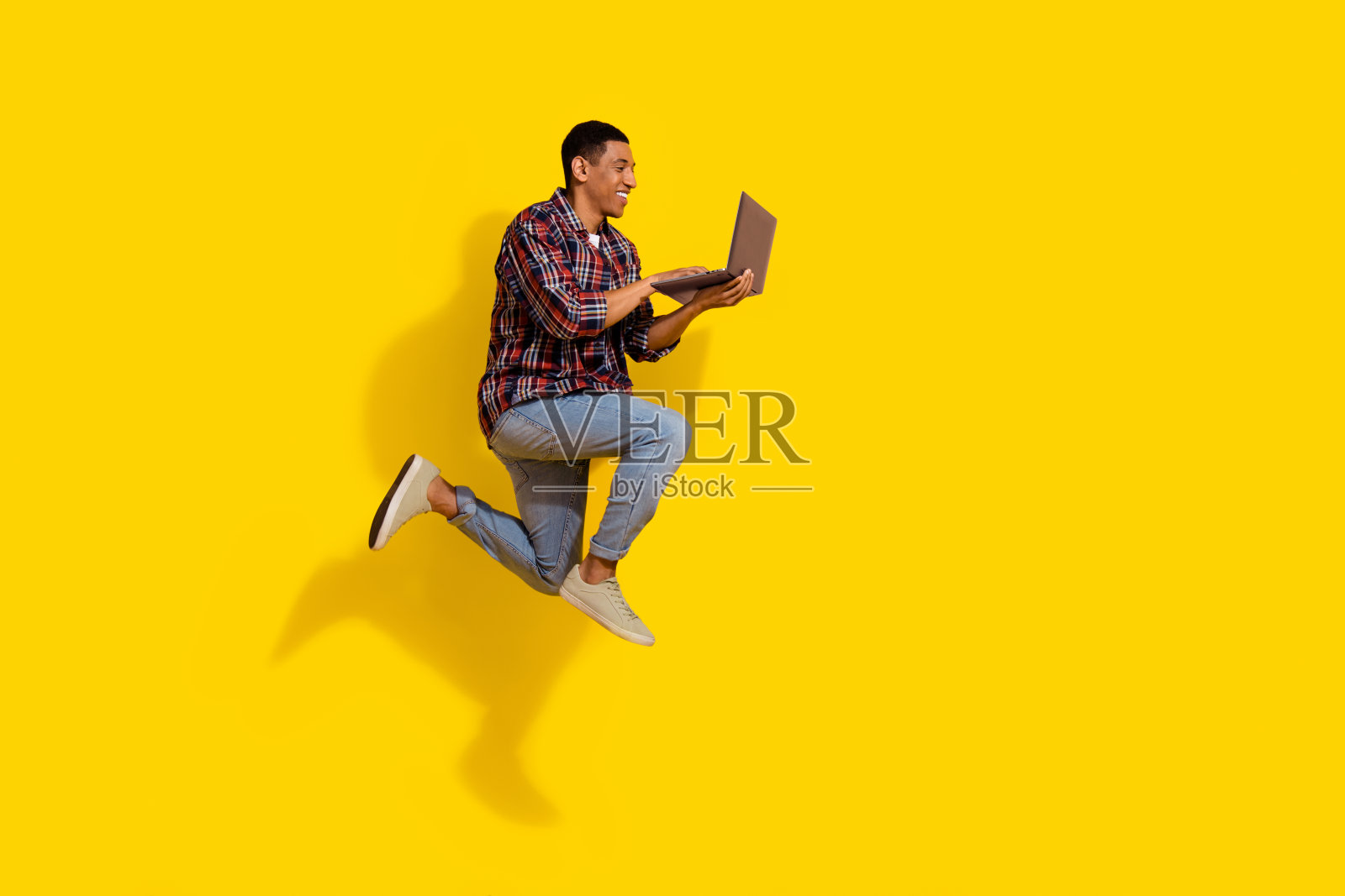 聪明的男人穿着格子衬衫牛仔裤在空旷的空间里飞翔，看着笔记本电脑孤立在鲜艳的黄色背景上照片摄影图片
