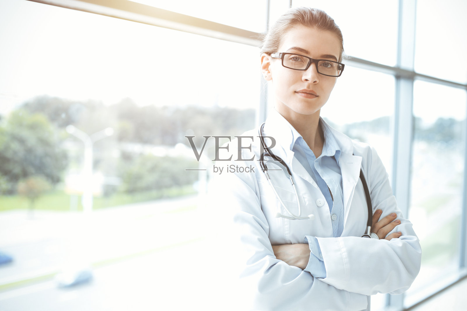 在阳光明媚的医院里，带着听诊器的美女医生微笑着，随时准备帮助病人。医学的概念照片摄影图片