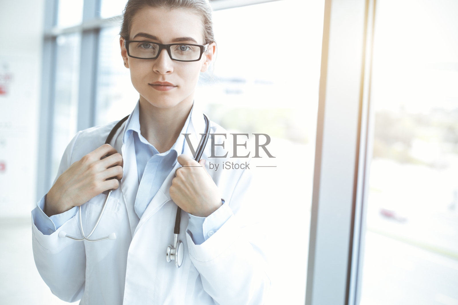 在阳光明媚的医院里，带着听诊器的美女医生微笑着，随时准备帮助病人。医学的概念照片摄影图片