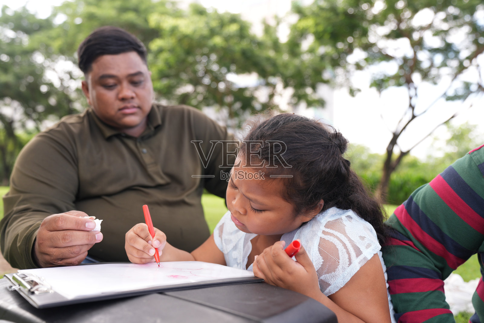 一个又大又胖的亚洲家庭，爸爸、妈妈、女儿在公园里野餐，他们坐着吃零食。他们坐着做作业，互相送花。他们很开心。照片摄影图片