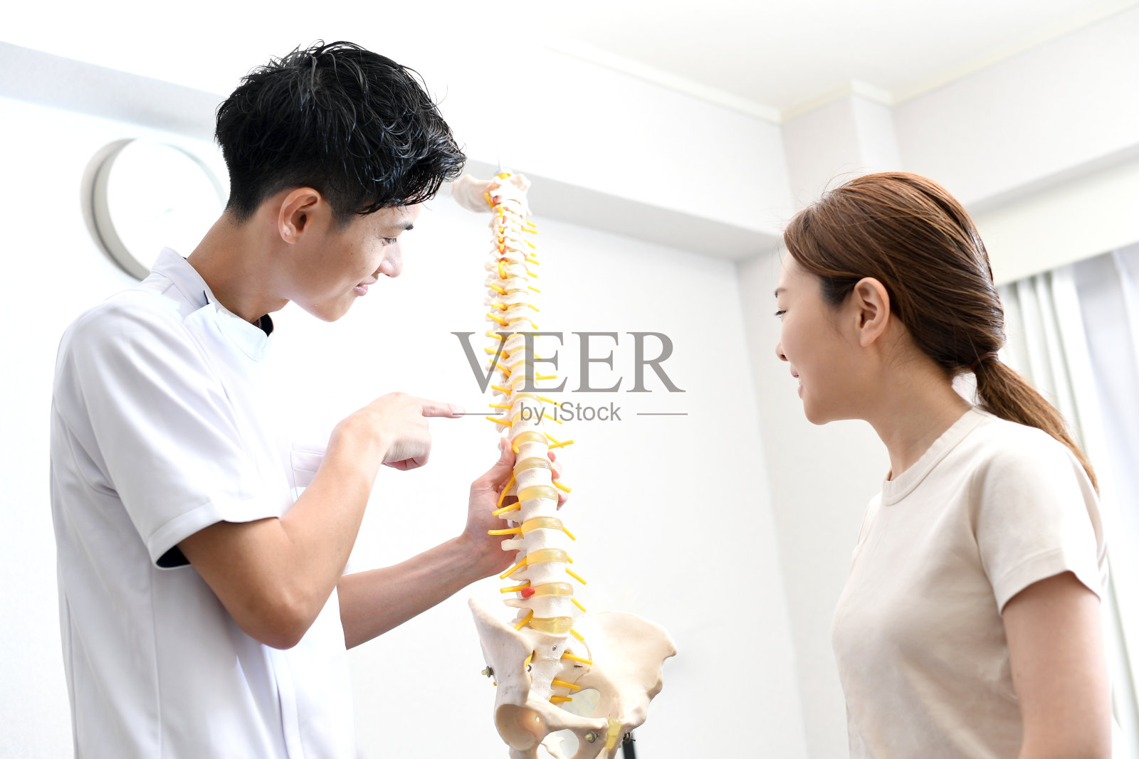 一位身穿白大褂的亚洲男性脊椎按摩师拿着脊椎模型向病人解释。照片摄影图片