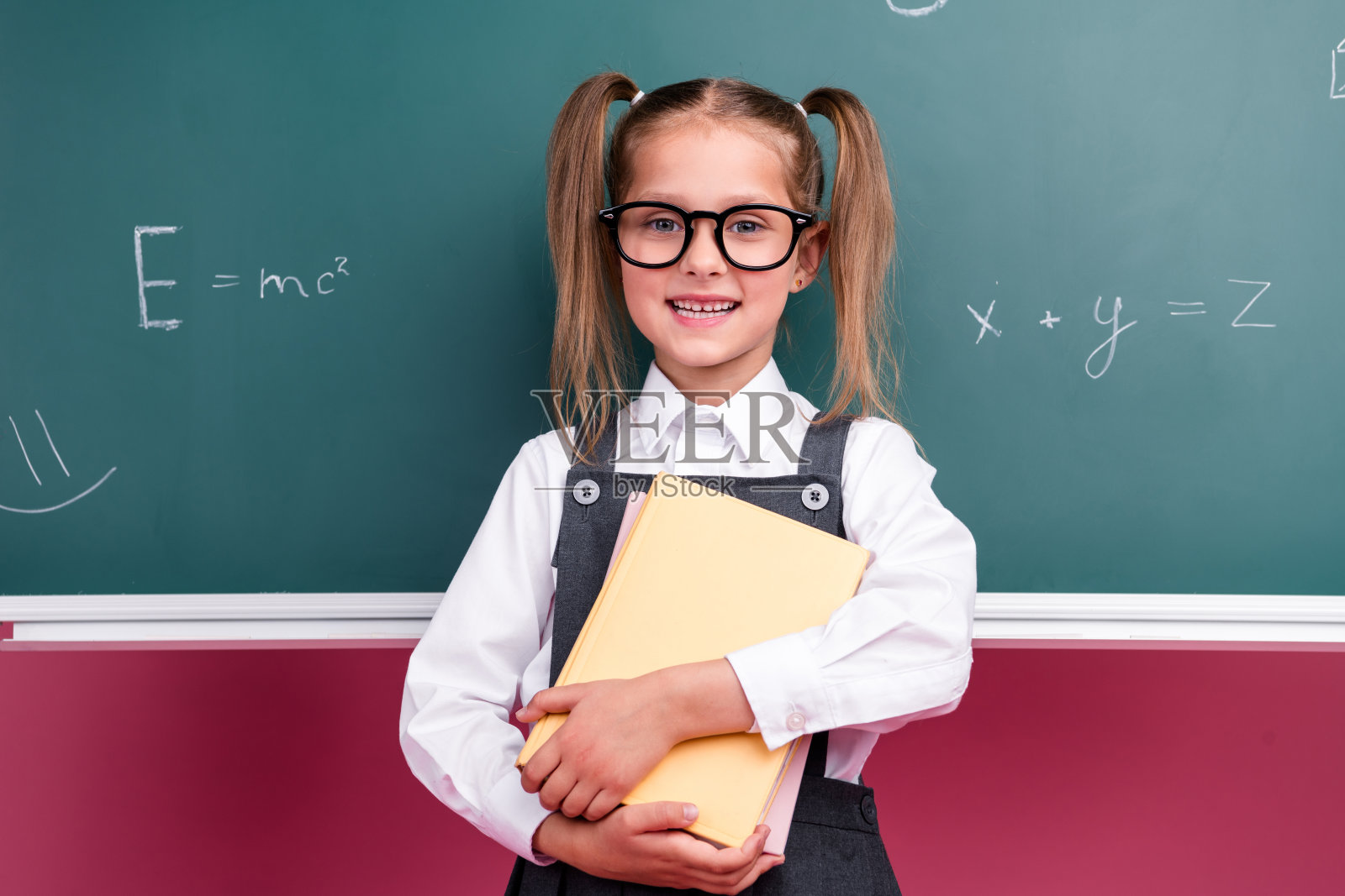 照片中可爱的小女生勤奋地拿着书本，穿着时髦的校服教室黑板背景照片摄影图片