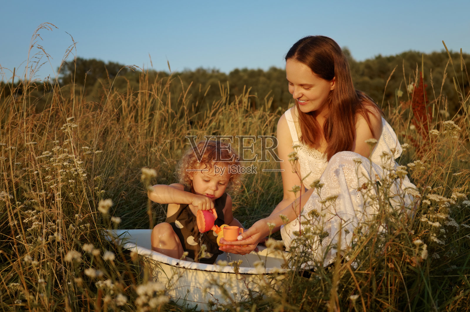 微笑的高加索妇女和波浪头发的孩子一起在五颜六色的草地上玩耍，在花盆里洗澡，享受夏日无忧无虑的时刻照片摄影图片