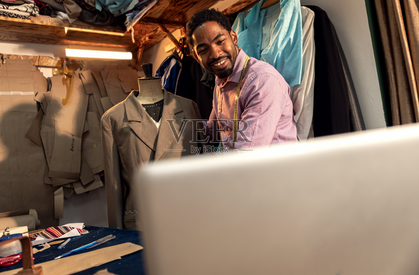 男时装裁缝通过笔记本电脑与客户视频通话，在他的工作室设计西装。照片摄影图片