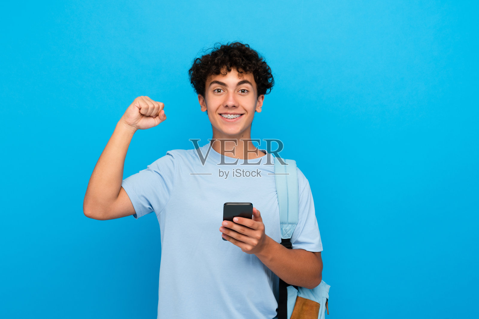 快乐的男孩背着背包，拿着智能手机，兴奋地做着获胜的手势，庆祝成功照片摄影图片