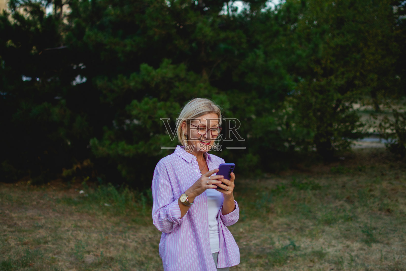 一个成熟的金发女人的肖像，戴着眼镜，穿着粉红色的衬衫，站在城市公园里，拿着手机照片摄影图片