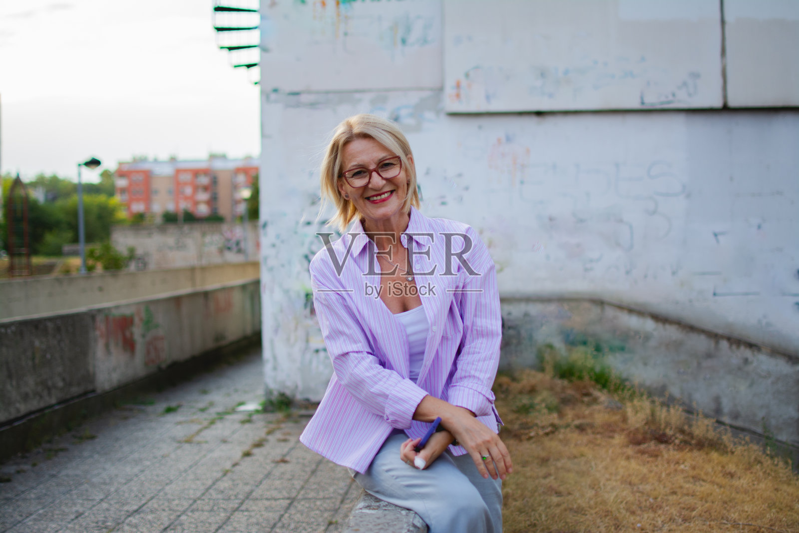 一个成熟的金发女人的肖像，戴着眼镜，穿着粉红色的衬衫，坐在公寓楼前照片摄影图片