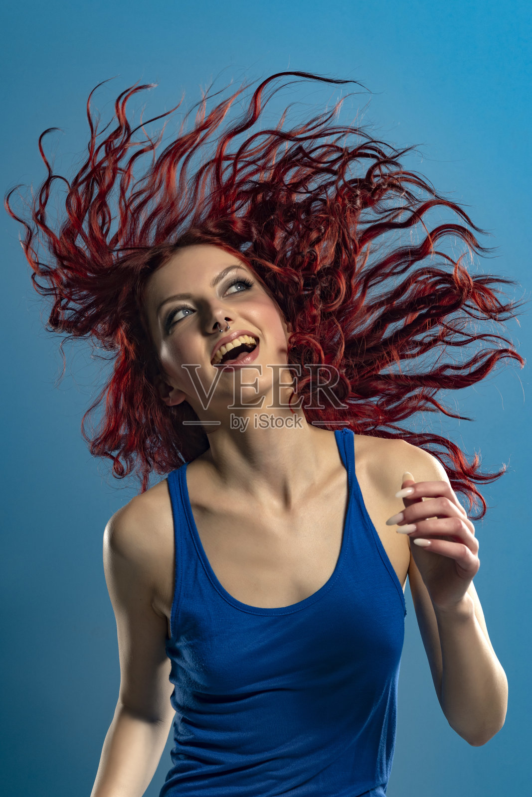 一个年轻快乐的跳舞女人的肖像，在蓝色的工作室背景上穿着紧身的蓝色连衣裙和长长的红色波浪头发照片摄影图片