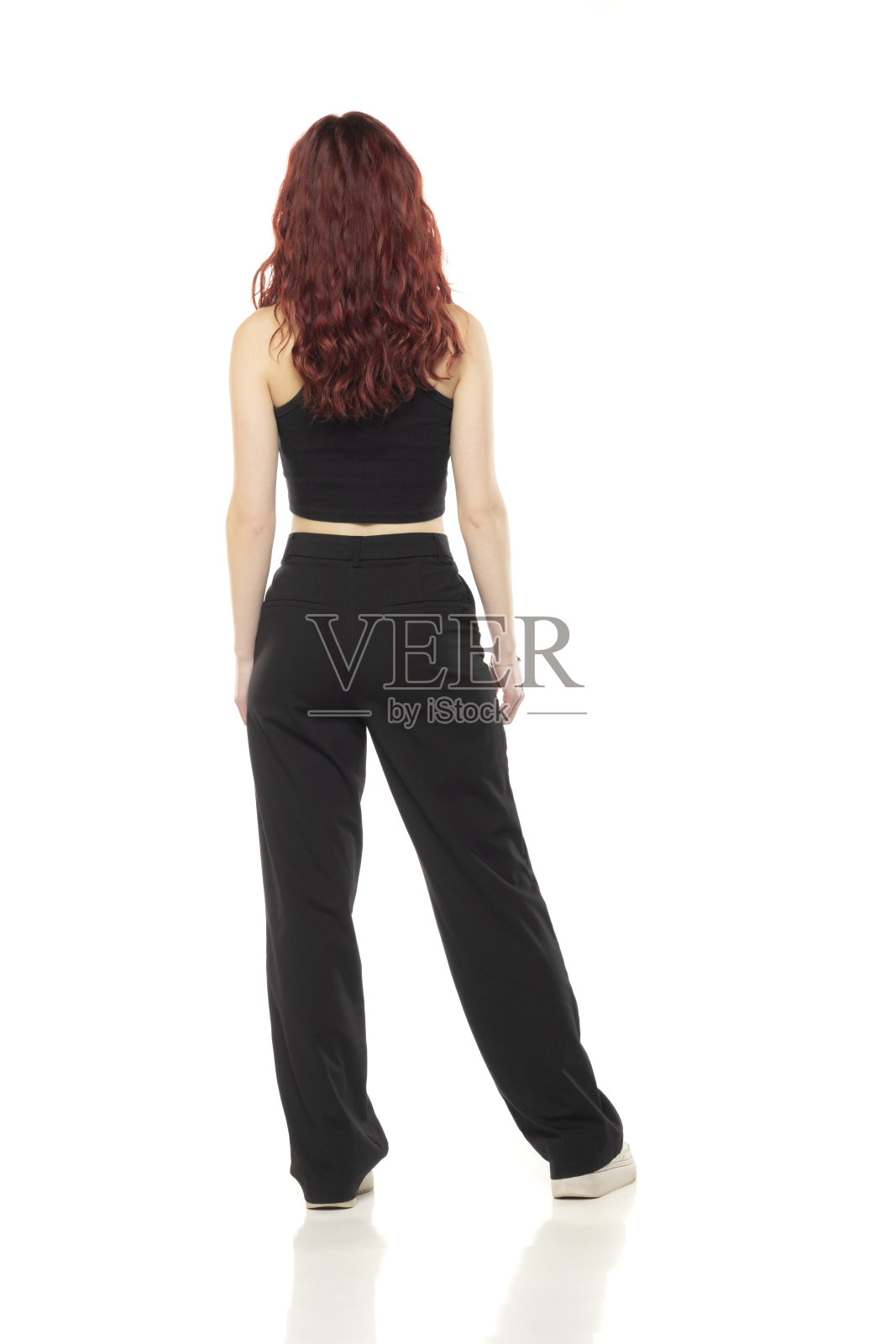 迷人的年轻红发女子在工作室里摆姿势，穿着黑色短衬衫和黑色裤子，背景是白色的。后视图照片摄影图片