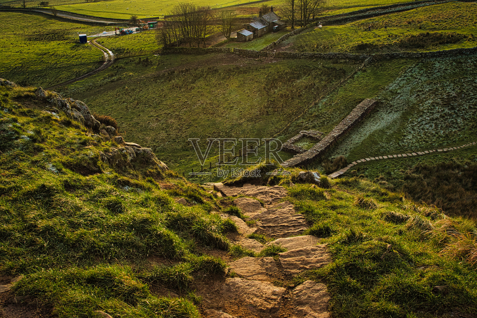 在英国诺森伯兰郡的西卡莫尔峡谷，日落时分，一条质朴的乡村小路穿过起伏的绿色山丘，远处有一个小村庄和蜿蜒的道路。照片摄影图片